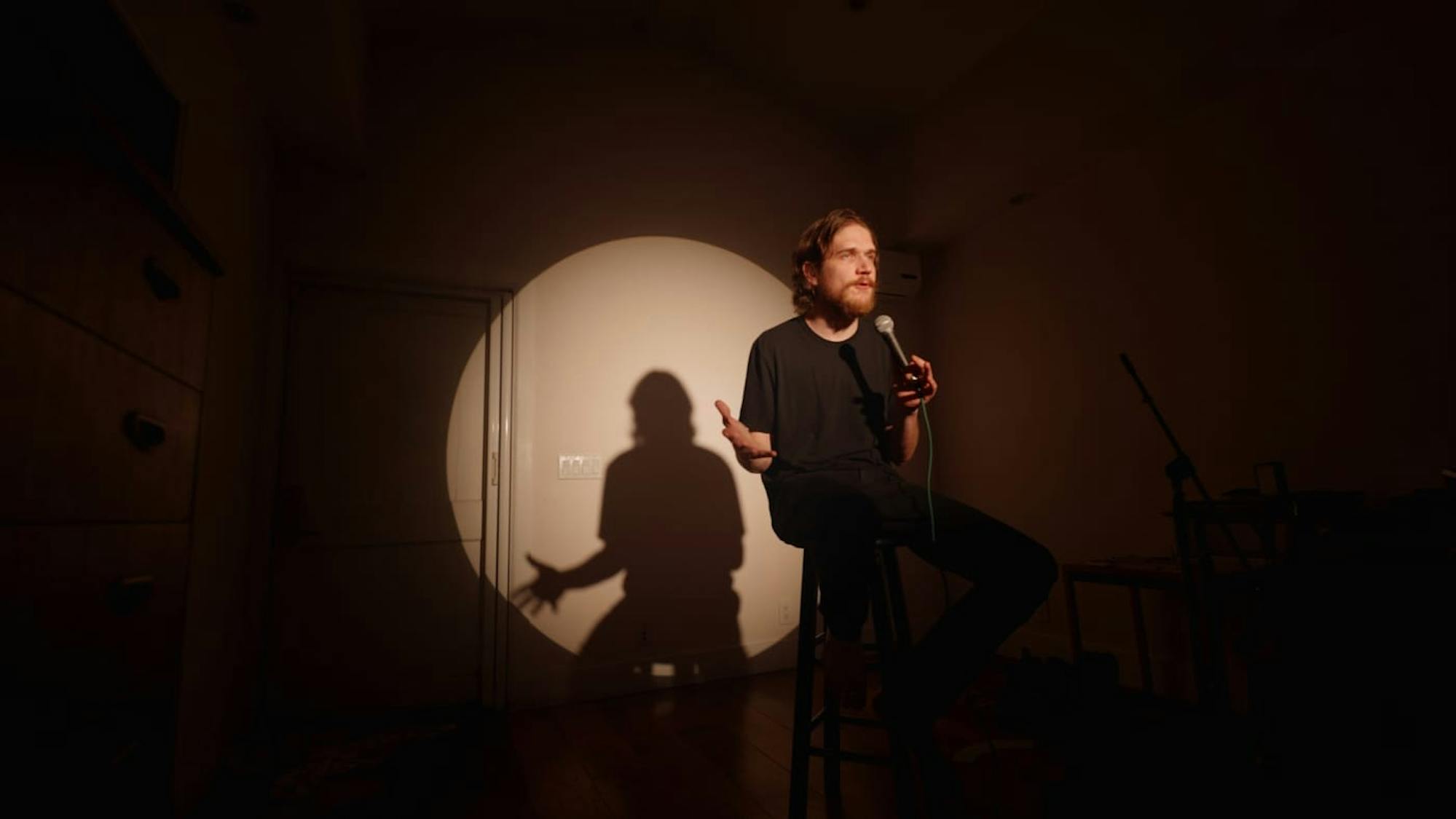 Bo Burnham sits on a stool with a single spotlight. He wears all black, and speaks into a microphone. Because the rest of the shot is dark it’s difficult to ascertain what else is in the room
