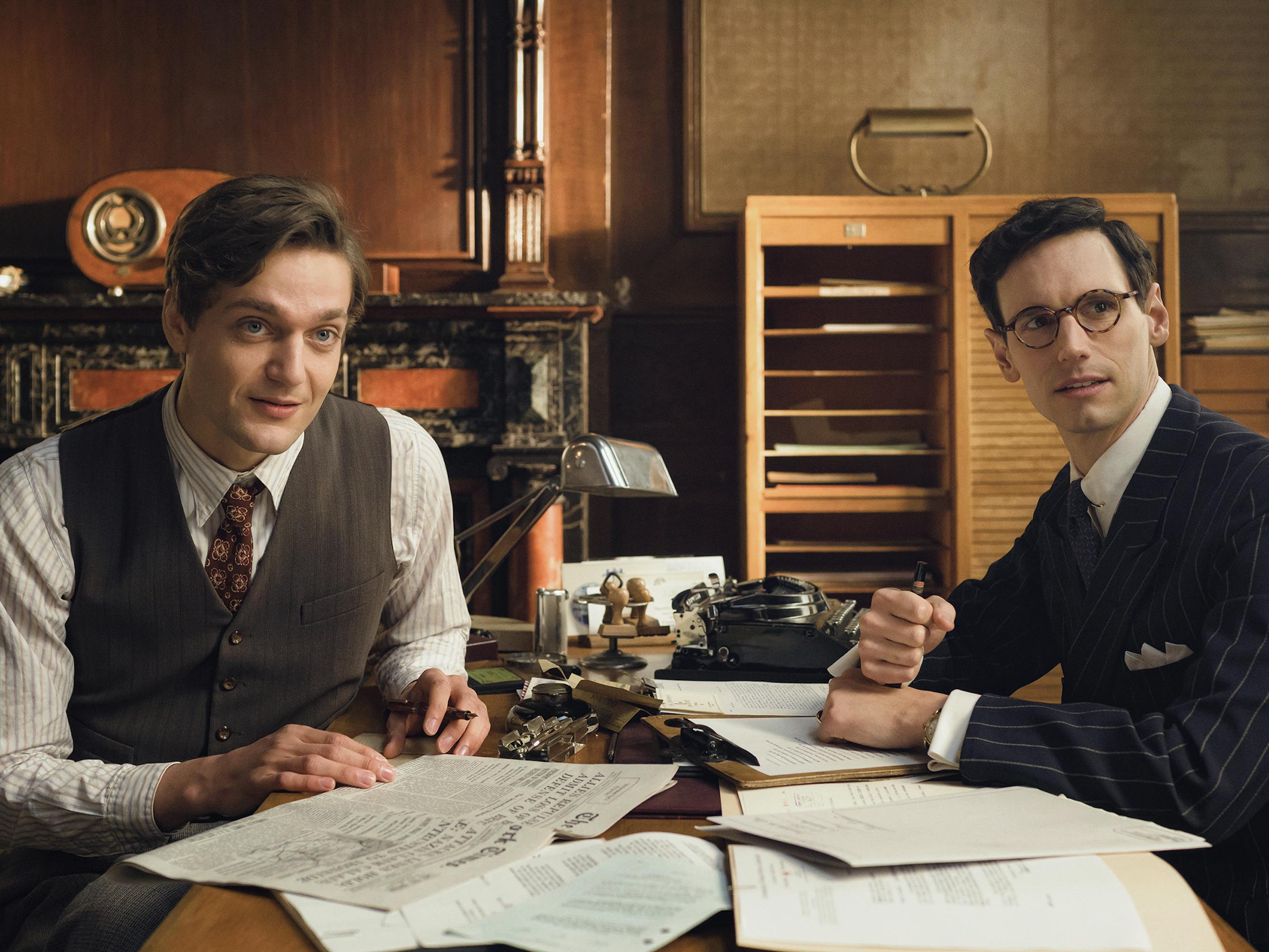 Albert Hirschman (Lucas Englander) and Varian Fry (Cory Michael Smith) sit at a desk reading through some papers looking at someone offscreen. Both wear sharp suits and look very prim.