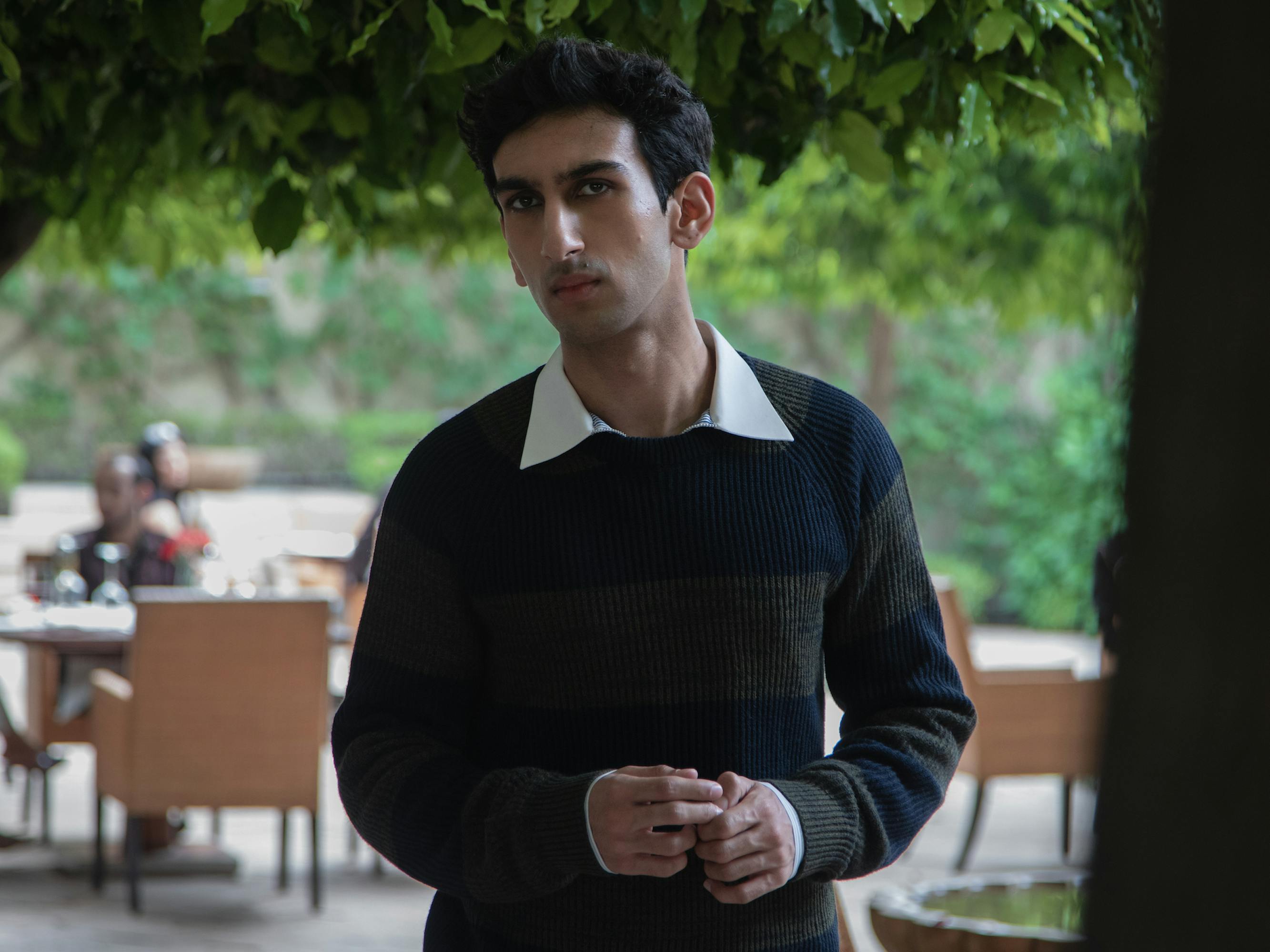 Sharan (Moses Koul) wears a navy and green striped sweater over a white collared shirt and looks dubious, standing in an outdoor area with brown tables and chairs. 