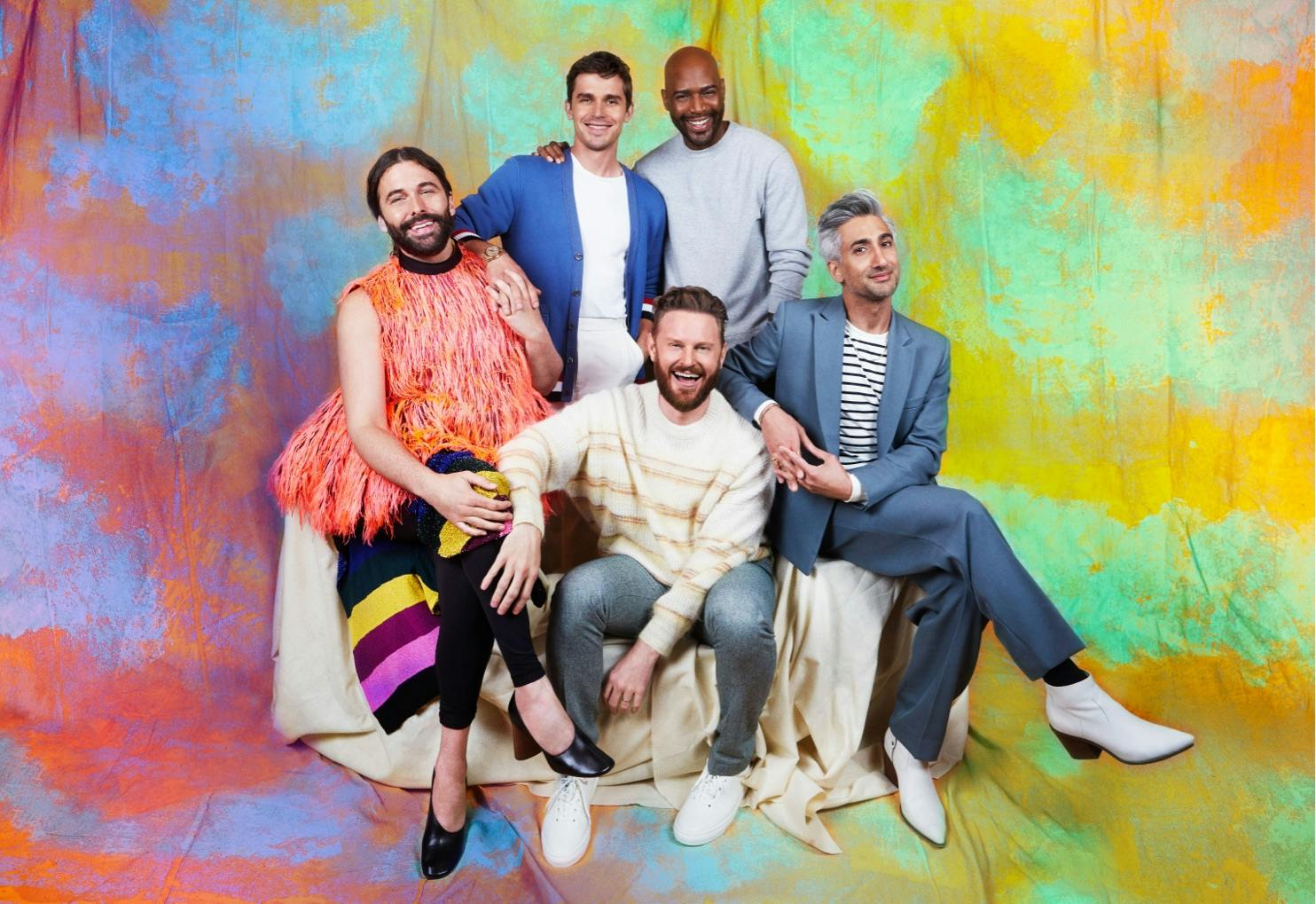 The Fab Five sit together against an orange and green spotted background. From left to right; Jonathan Van Ness wears a furry orange vest, Antoni Porowski wears a blue cardigan, Karamo Brown wears a light blue sweater, Bobby Berk wears jeans and a striped sweater, Tan France wears a grey suit with white boots.