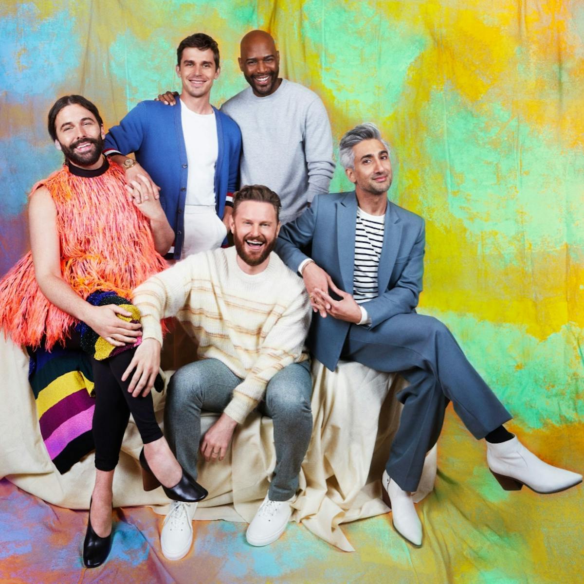 The Fab Five sit together against an orange and green spotted background. From left to right; Jonathan Van Ness wears a furry orange vest, Antoni Porowski wears a blue cardigan, Karamo Brown wears a light blue sweater, Bobby Berk wears jeans and a striped sweater, Tan France wears a grey suit with white boots.