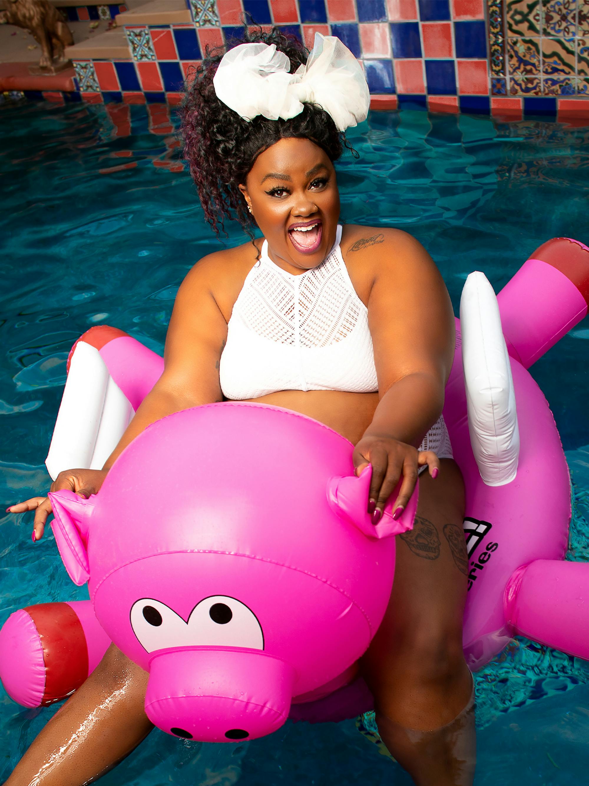 Nicole Byer sits on an inflatable pig, floating in a pool.