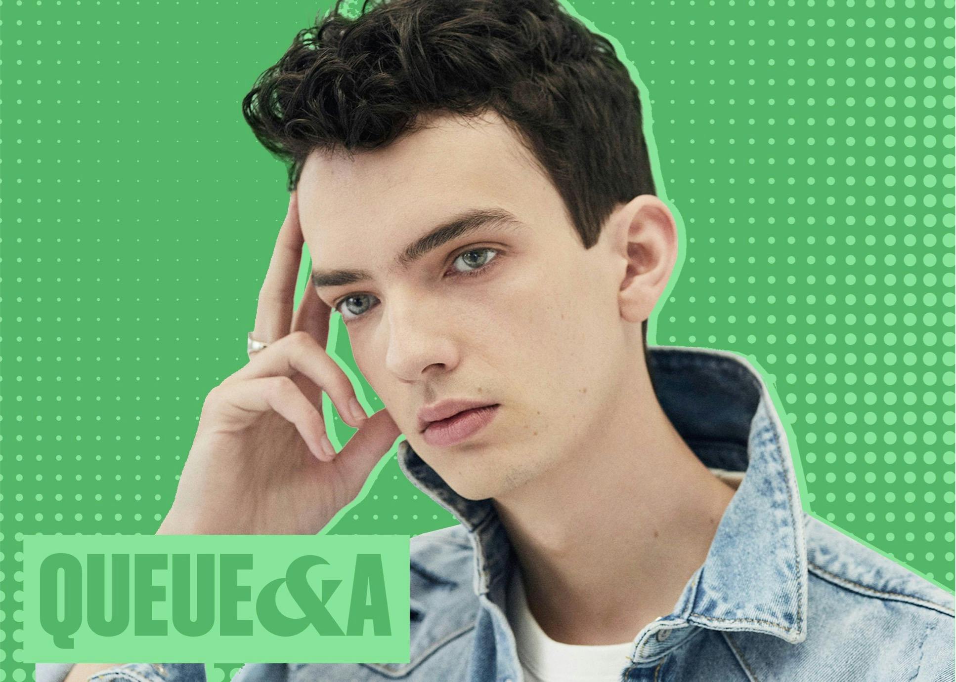 Kodi Smit-McPhee wears a denim shirt with a white t-shirt underneath. He raises his fingers to his temple. The background is green speckled with lighter green and in the bottom left hand corner reads: Queue &A.