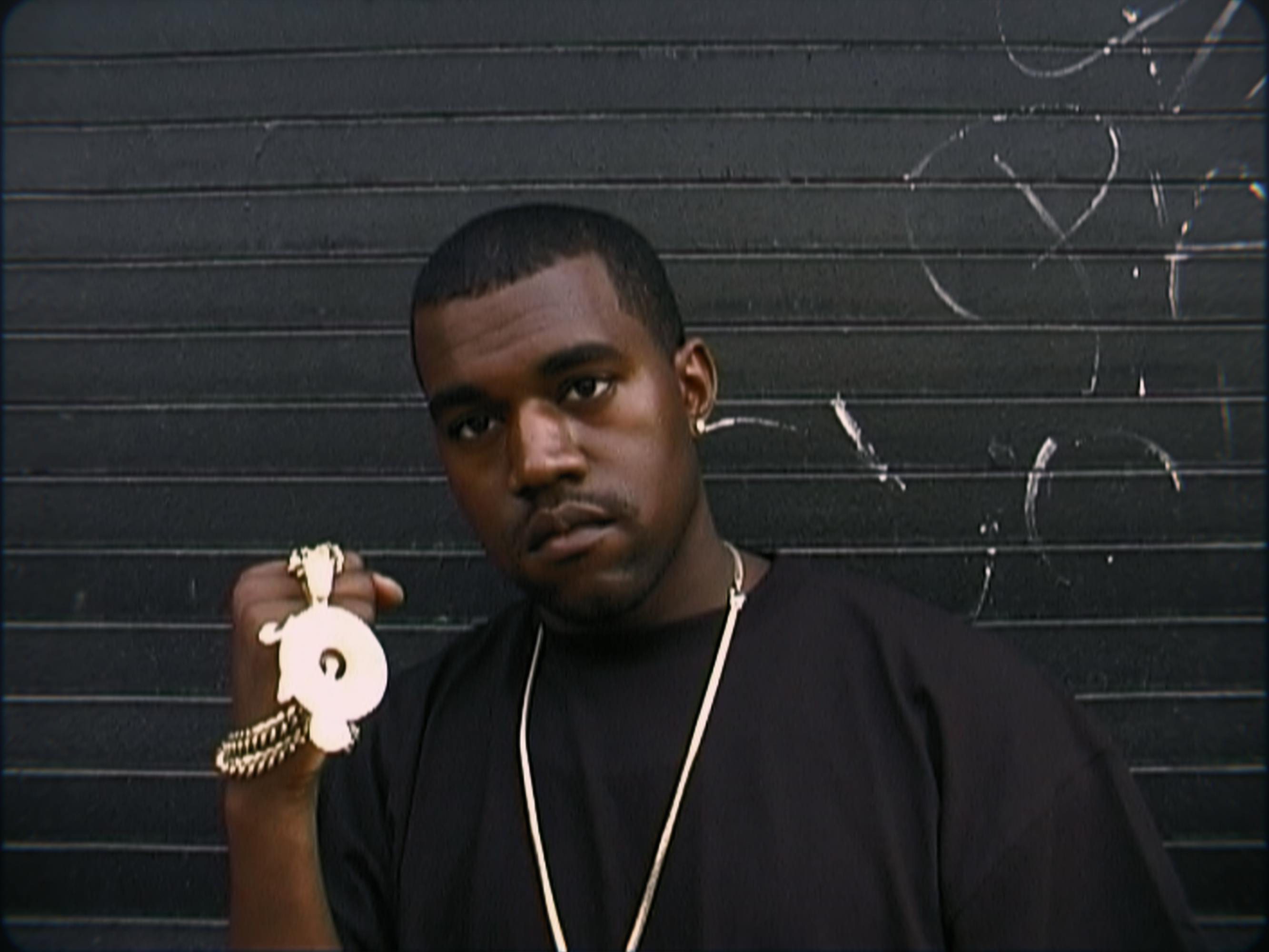 Kanye West holds a gold necklace, and wears a long chain and navy shirt. 