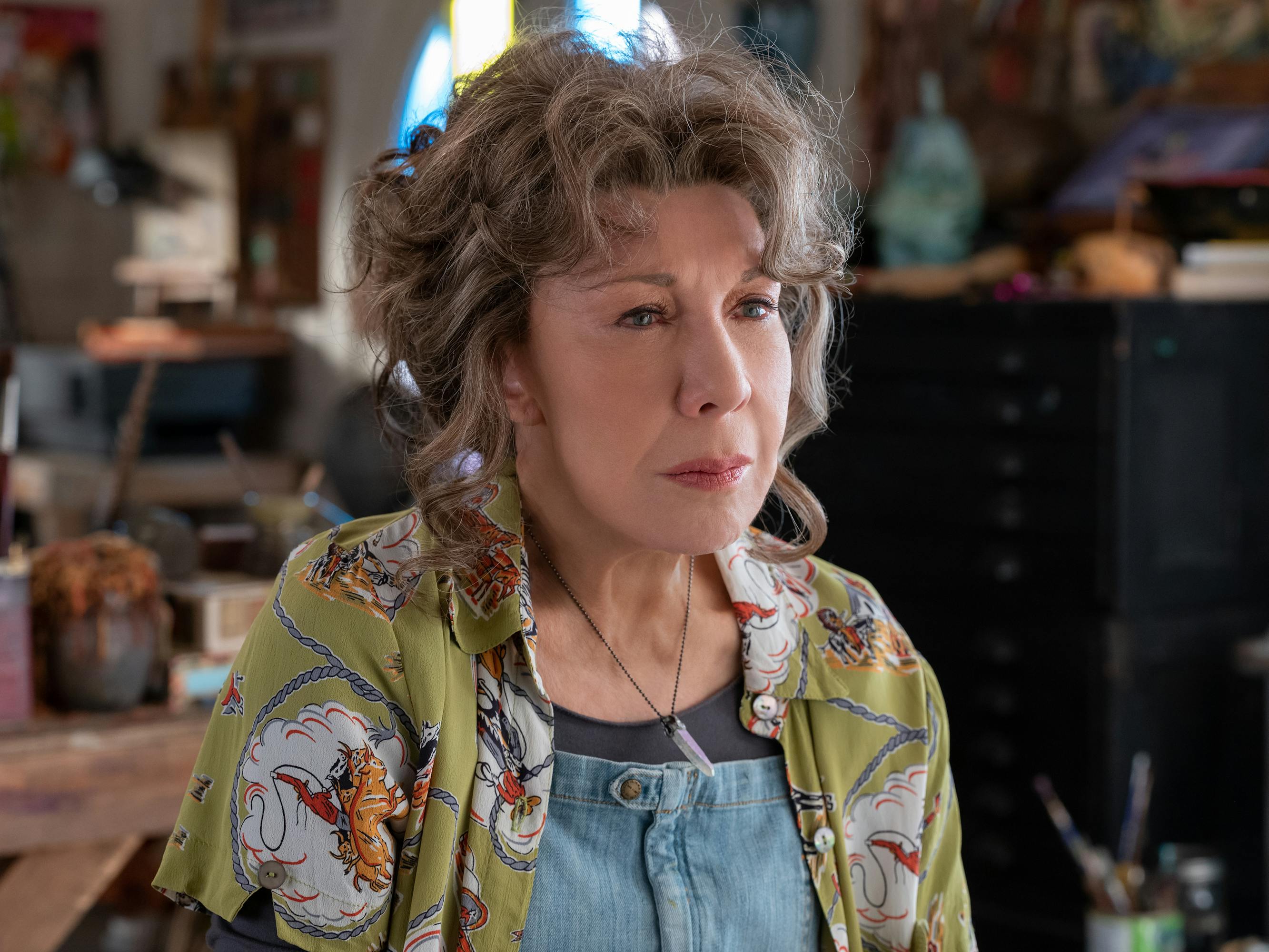 Lily Tomlin wears a green flowered shirt and jean overalls.