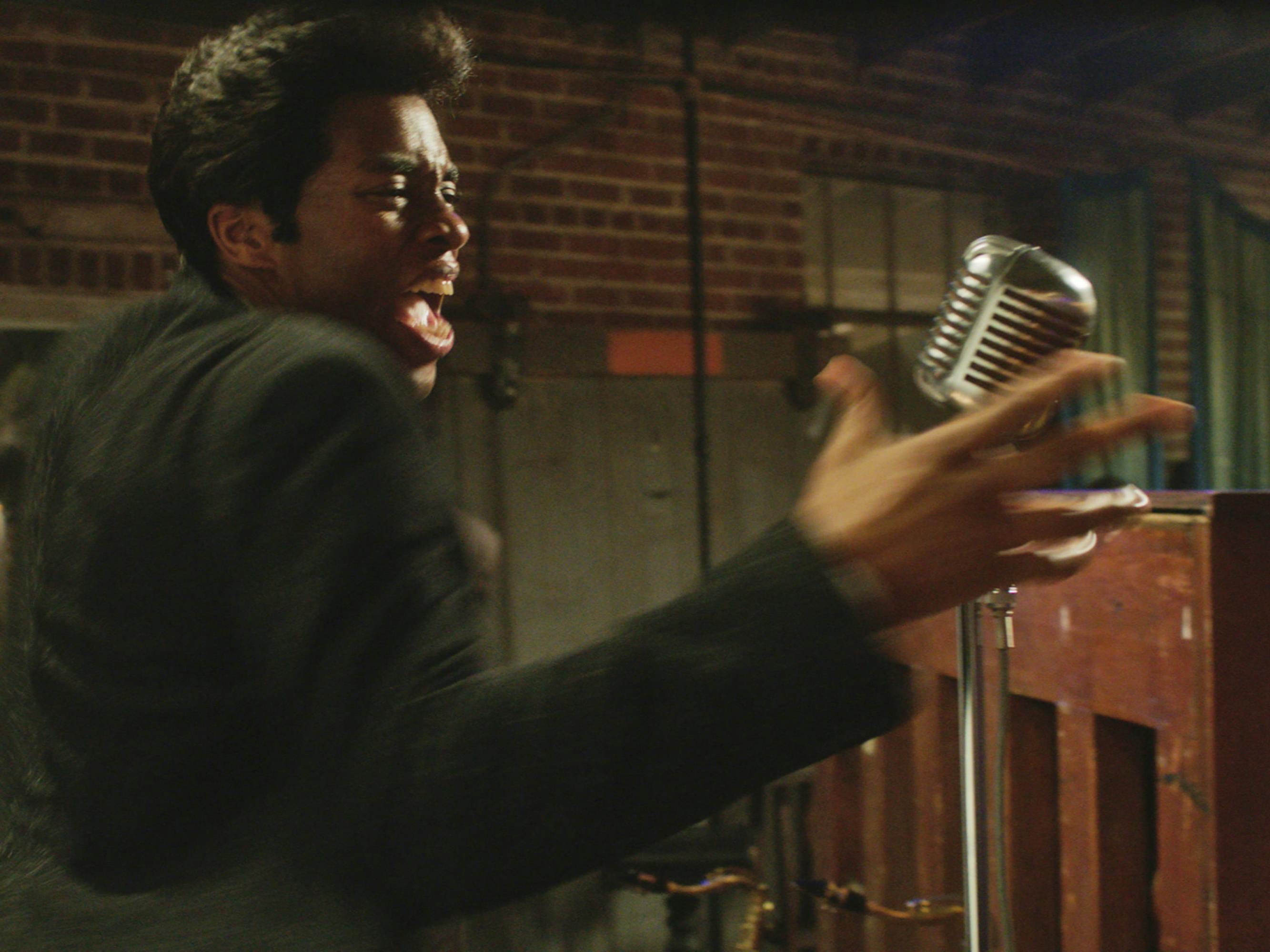 Boseman as James Brown. He wears a black suit and his hair in Brown’s signature conk pompadour. His mouth is open wide, and he reaches for the microphone. Despite the dynamism of the shot, he appears to be in a practice space with brick walls, and other musical equipment strewn about. 