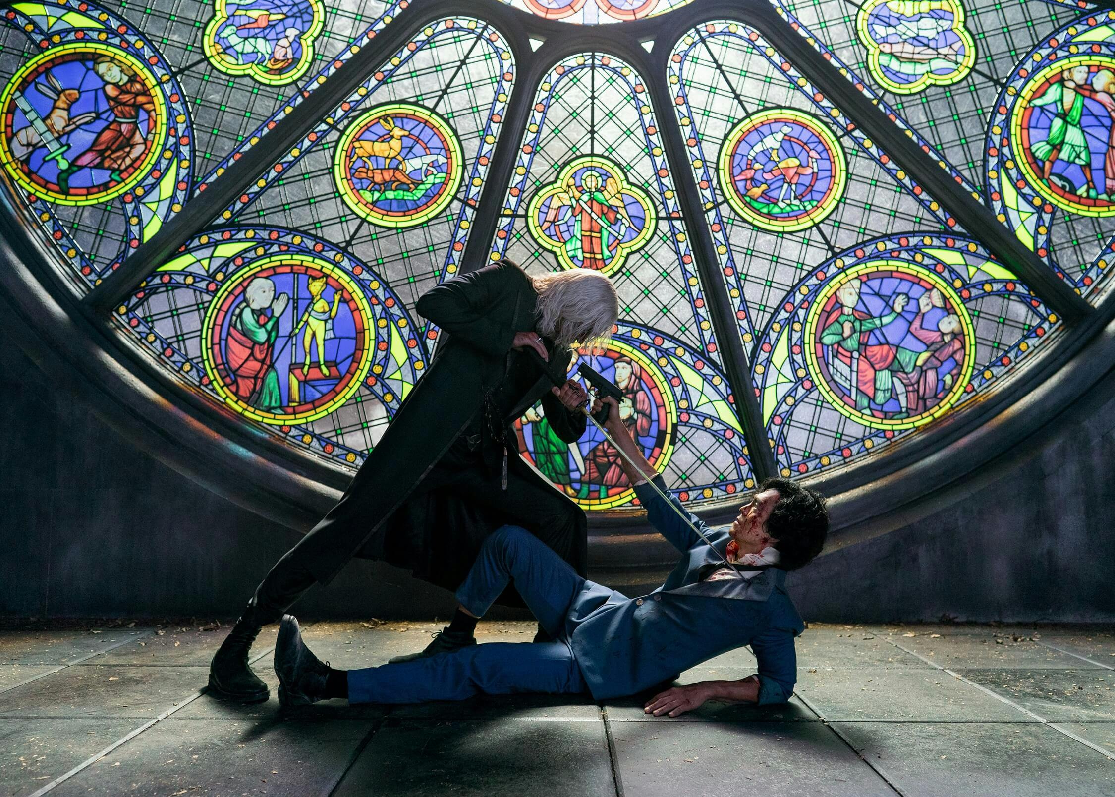 Alex Hassell and John Cho face off in front of a stained class window.