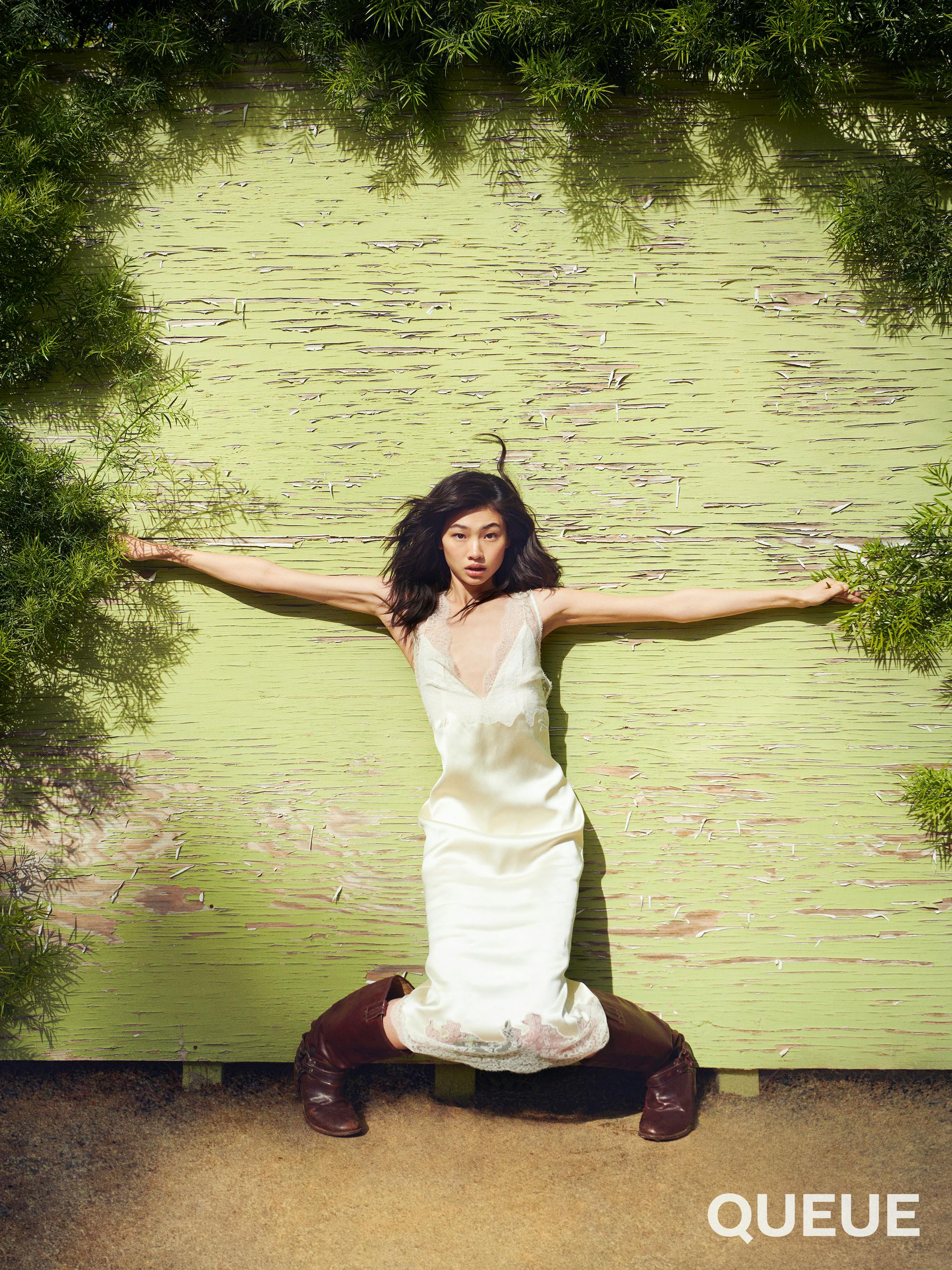 Jung Ho-yeon wears a white slip and brown boots and rests against a green wall.  