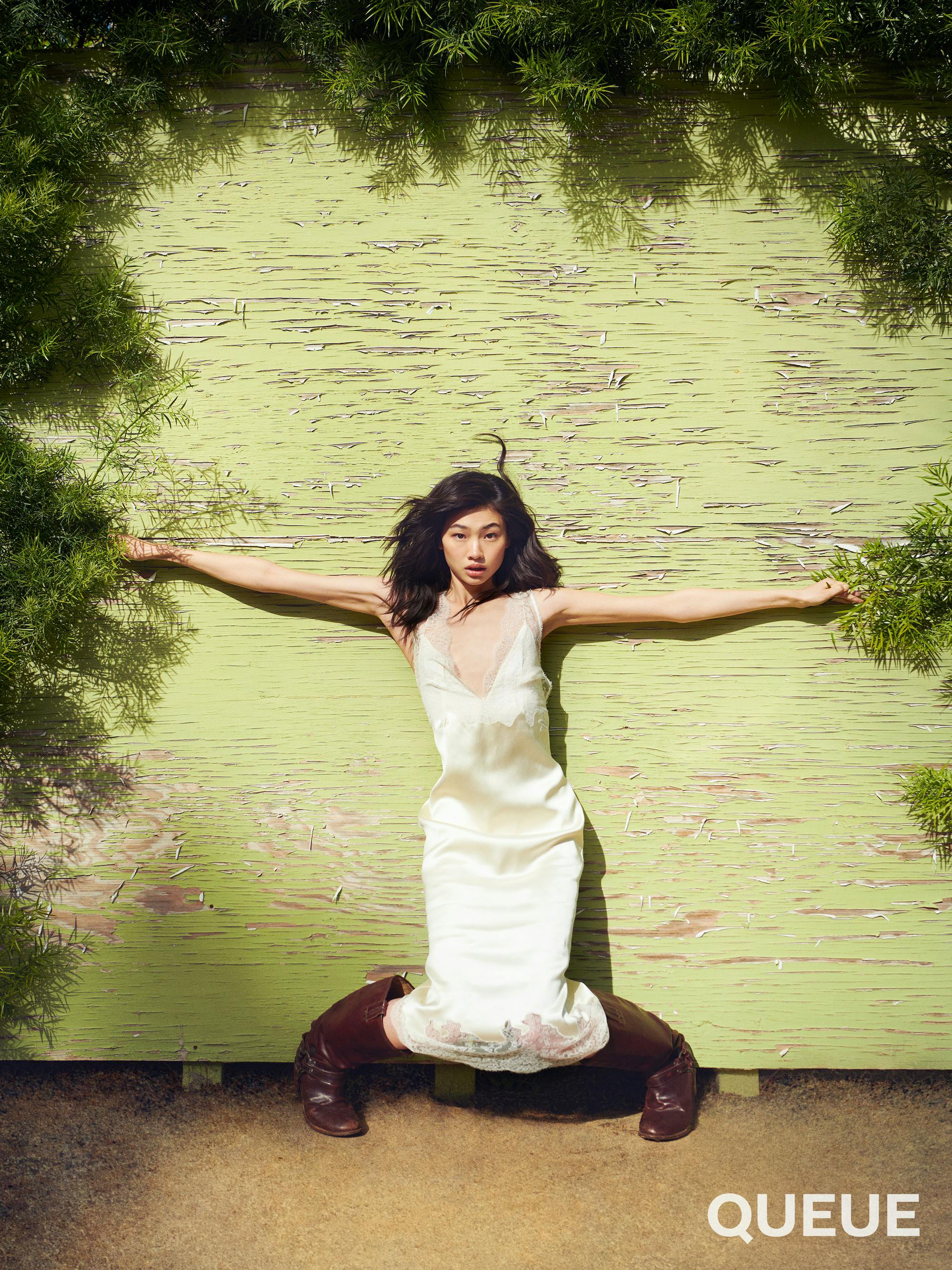 Jung Ho-yeon wears a white slip and brown boots and rests against a green wall.