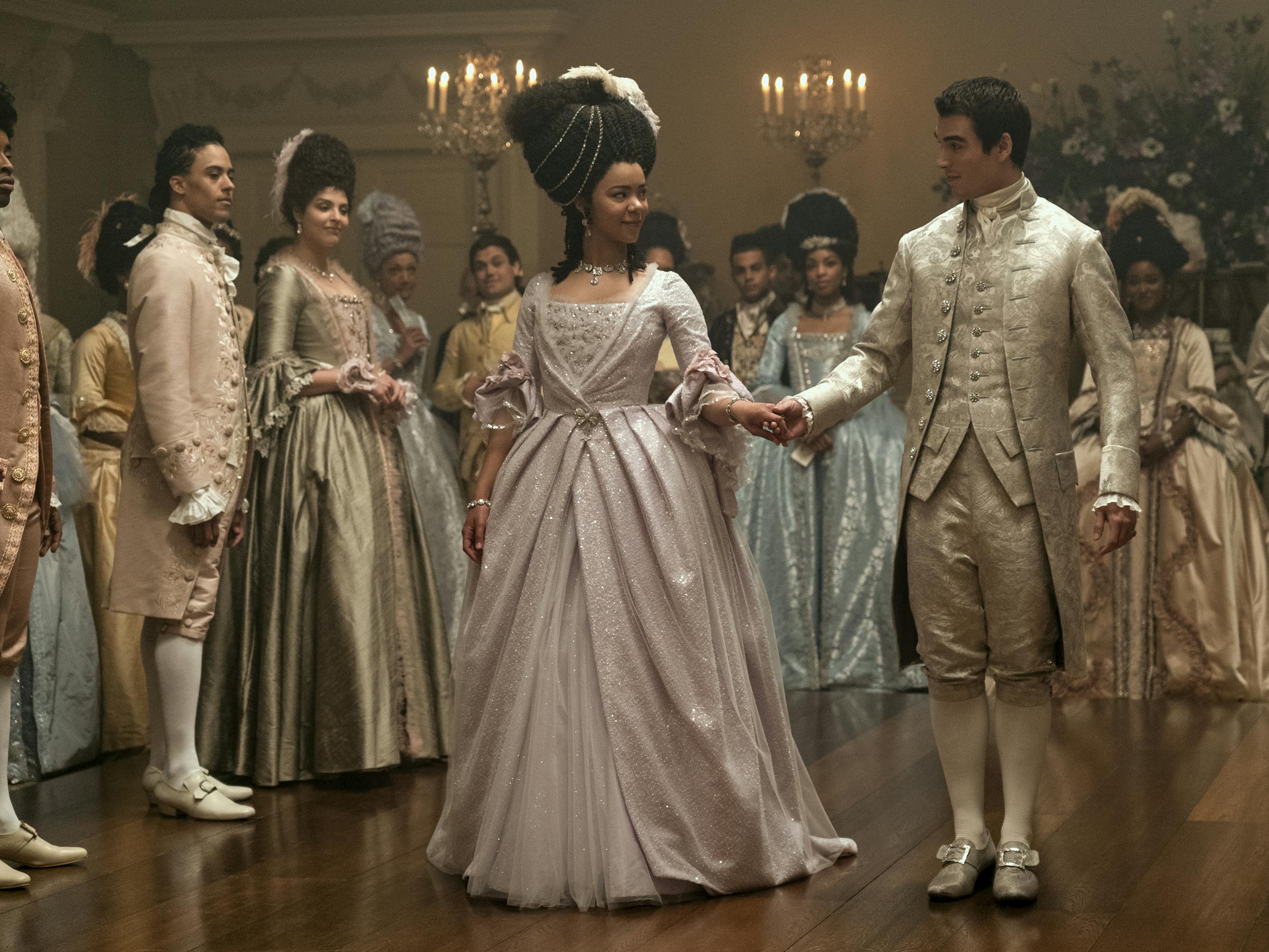 Queen Charlotte (India Amarteifio) and King George (Corey Mylchreest) dance together on a chandelier-lit dance floor, accompanied by some pastel-wearing people.