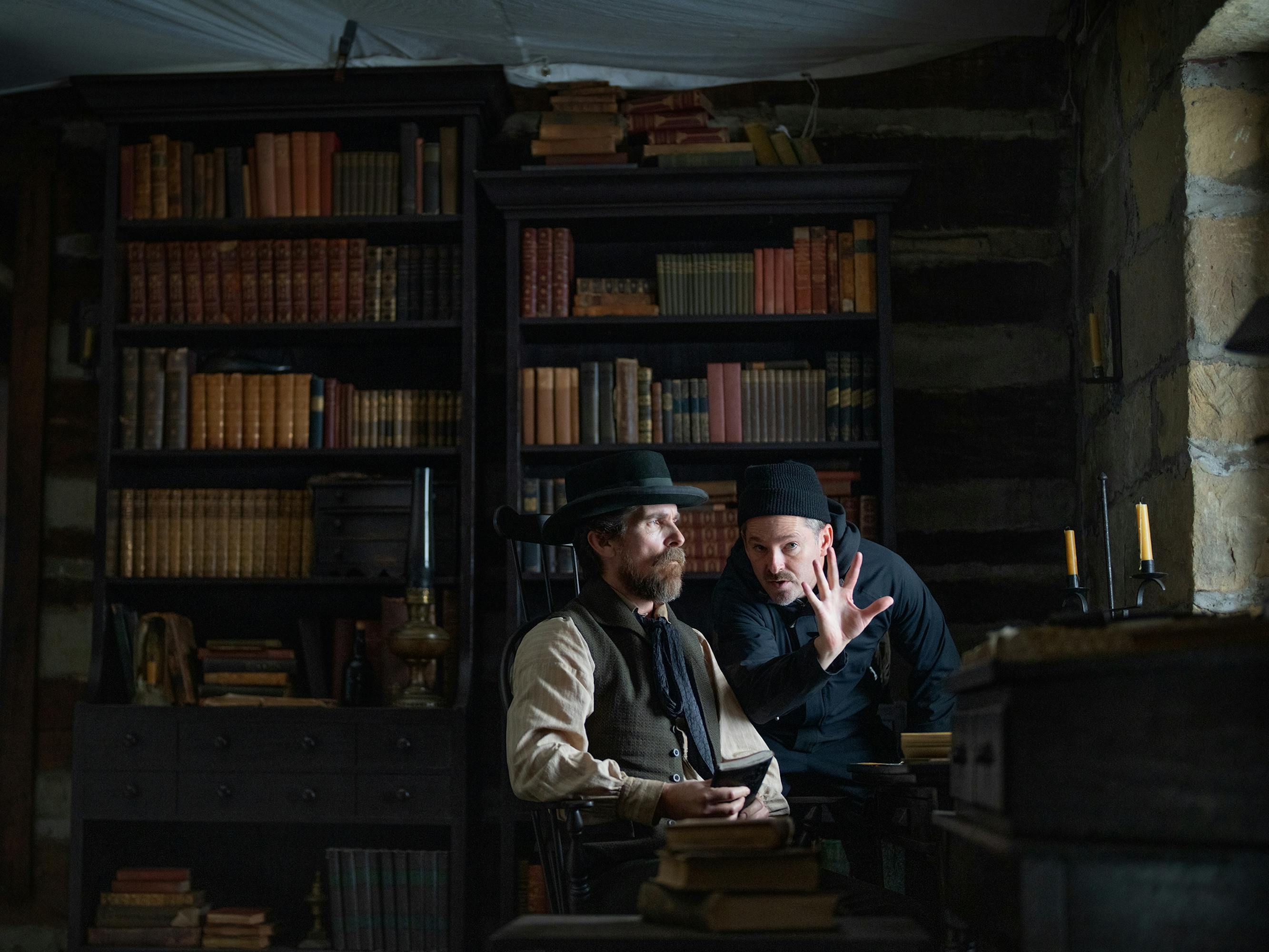 Christian Bale and Scott Cooper sit in a dark library together, with a bookshelf filled with old books behind them.