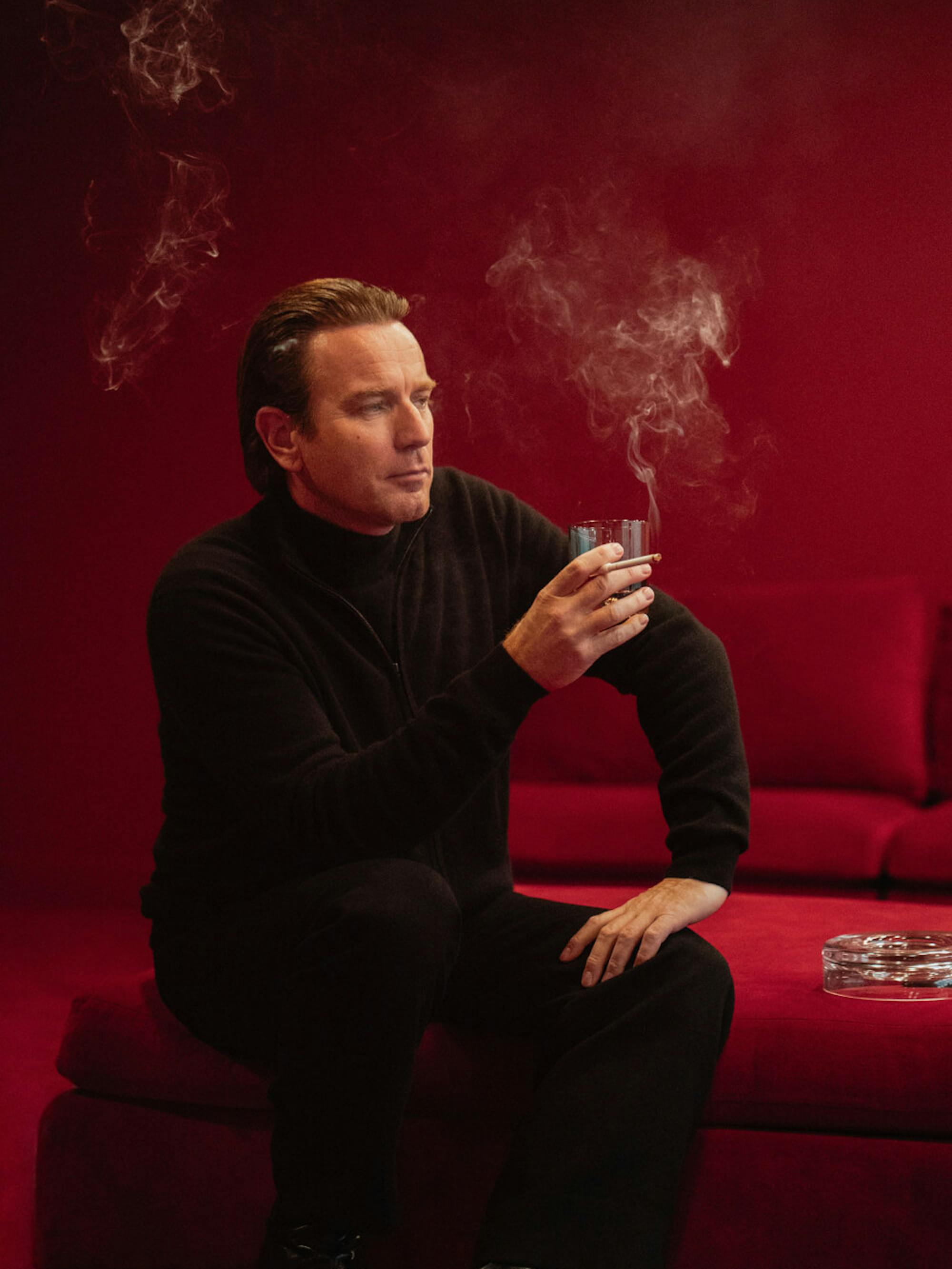 Halston wears his classic black ensemble as he smokes and drinks in a lush, red, velvet room.