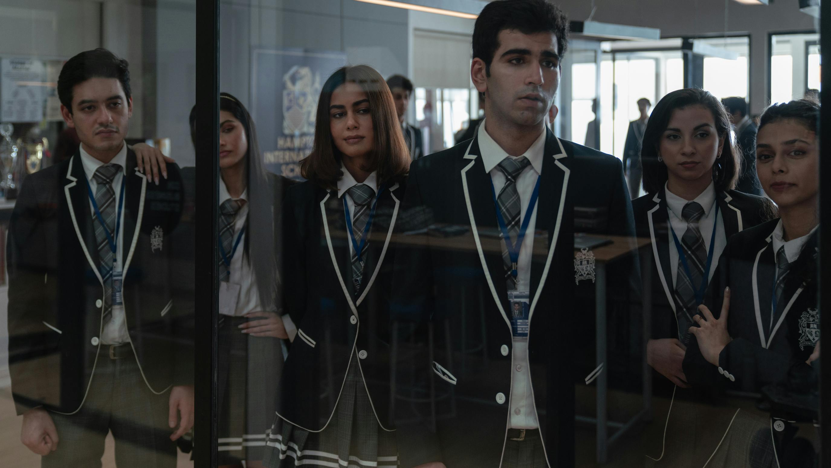 A line of students looks through a glass window. They all wear uniforms: grey skirts, grey pants, navy blazers, white shirts, blue lanyards, and plaid ties. 