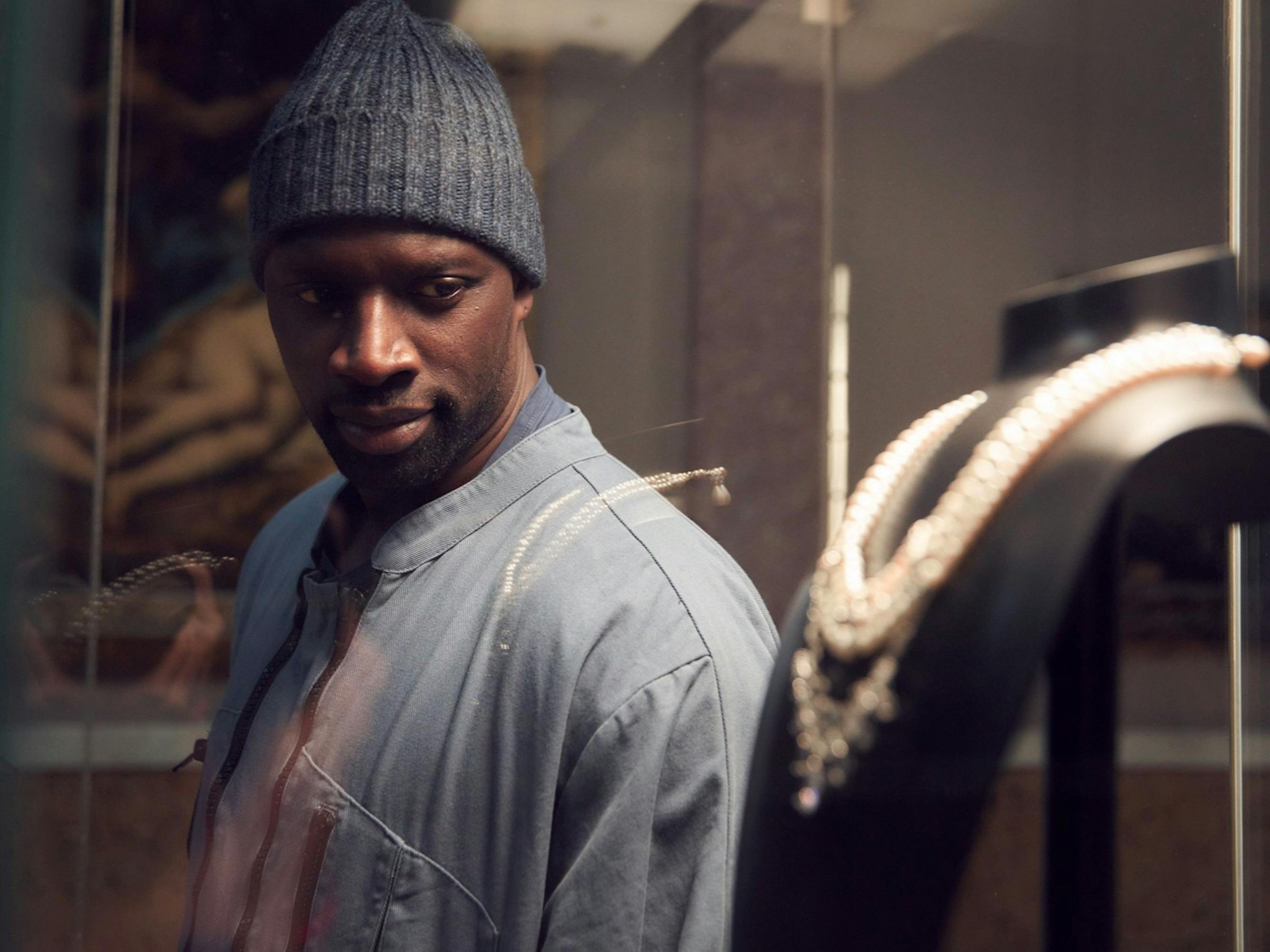 Assane Diop (Omar Sy) looks at a necklace on a black mannequin enclosed in a glass box. Sy wears a grey jacket and wool hat.