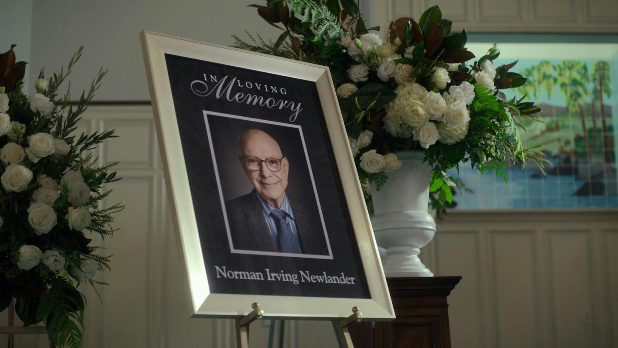 A portrait of Norman Newlander (Alan Arkin) sits propped up beside two vases of flowers. The scene appears to be a place of worship in which a funeral is taking place. The portrait is framed with gold and is predominately dark save the white lettering (“In Loving Memory of Norman Irving Newlander”). Newlander wears glasses, a blue shirt, navy tie, and navy blazer, and he smiles.