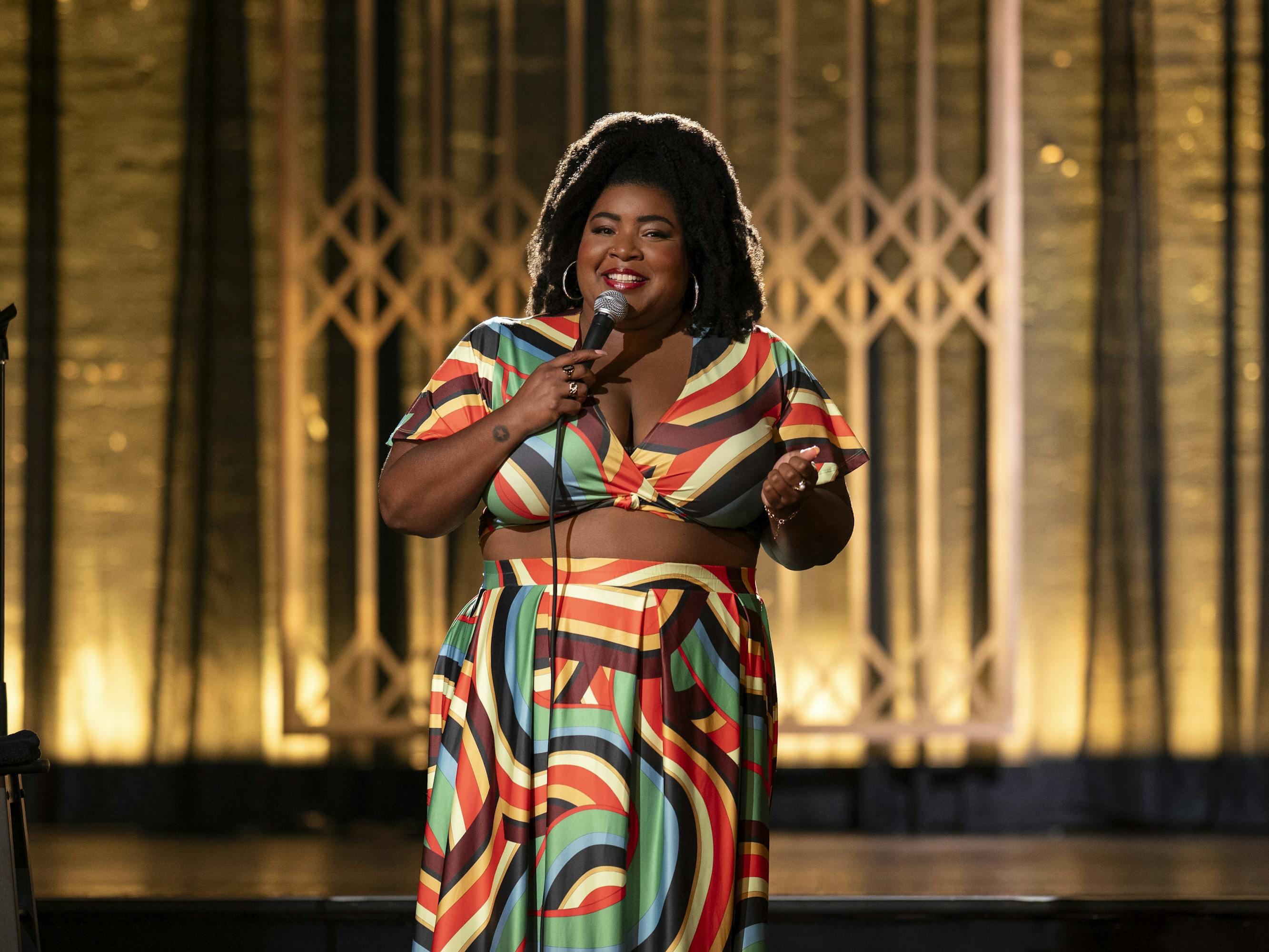 Dulcé Sloan in Verified Stand-Up wears a patterned skirt-and-top set and stands on an art-deco stage.