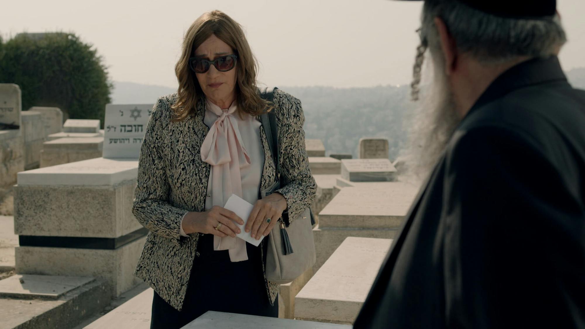 Nechama Yoktan and Shulem Shtisel stand in a cemetery. Nechama Yoktan looks upset even behind her large shades. She also wears a white shirt, pink scarf, and textured blazer. Shulem is in all black, and stands with his back to the viewer. The grim setting of the graveyard is offset by the bright sunlight and views behind them.