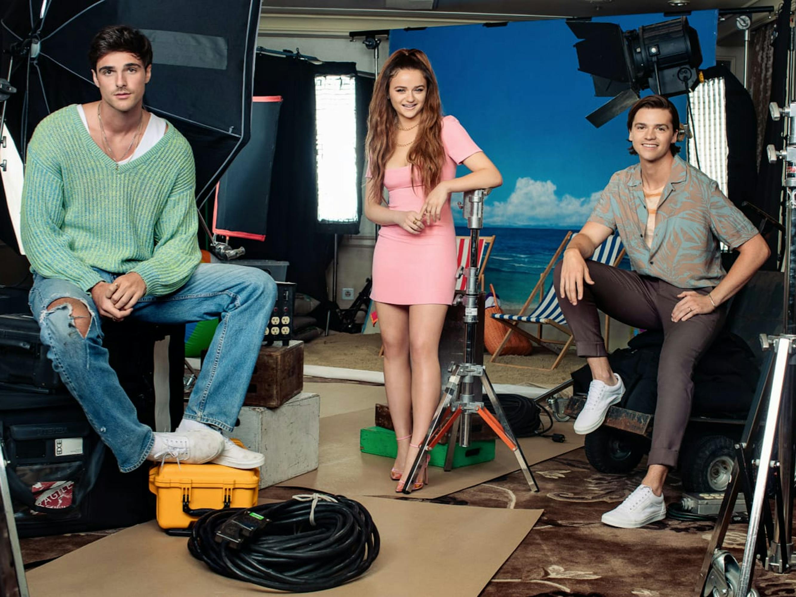 Jacob Elordi, Joey King, and Joel Courtney on set of The Kissing Booth. Elordi wears a green sweater and jeans, King wears a pink dress, and Courtney wears dark pants and a patterned shirt.