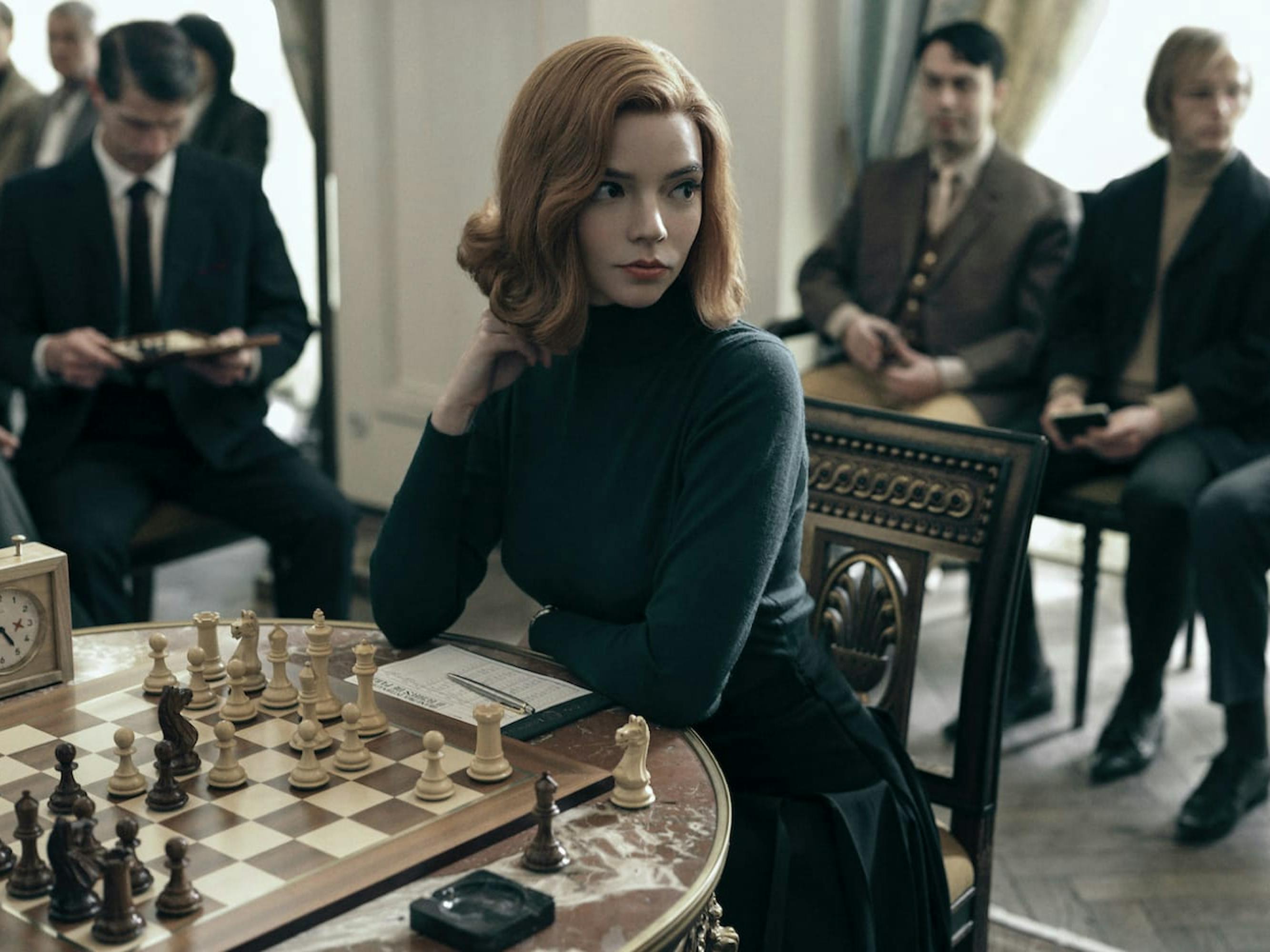 Beth Harmon (Anya Taylor-Joy) sits in front of a chess board at a competition, surrounded by men in dark suits. She also wears all black.