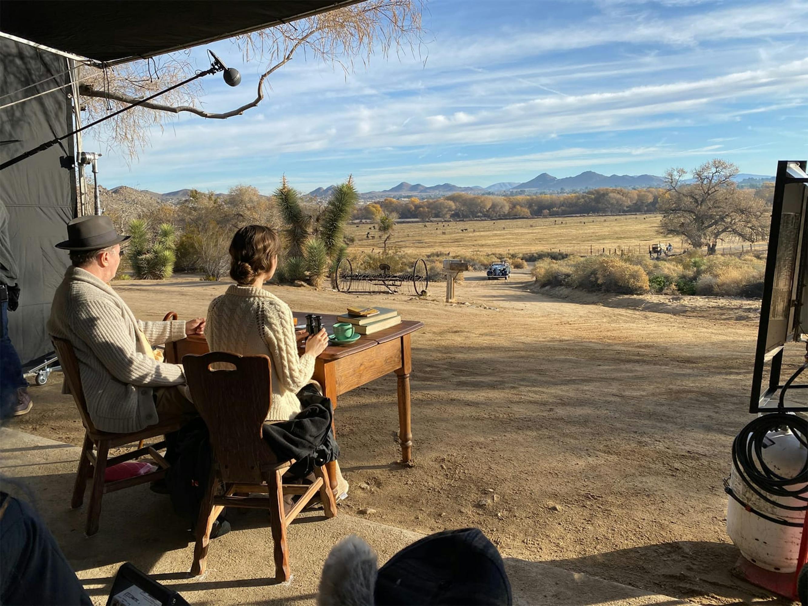 Oldman and Lily Collins in character on the set of Mank. The pair are seated at a table, gazing out at the backdrop that serves as the California desert. The sky is a bright blue and the expanse in front of them is a dusty yellow.
