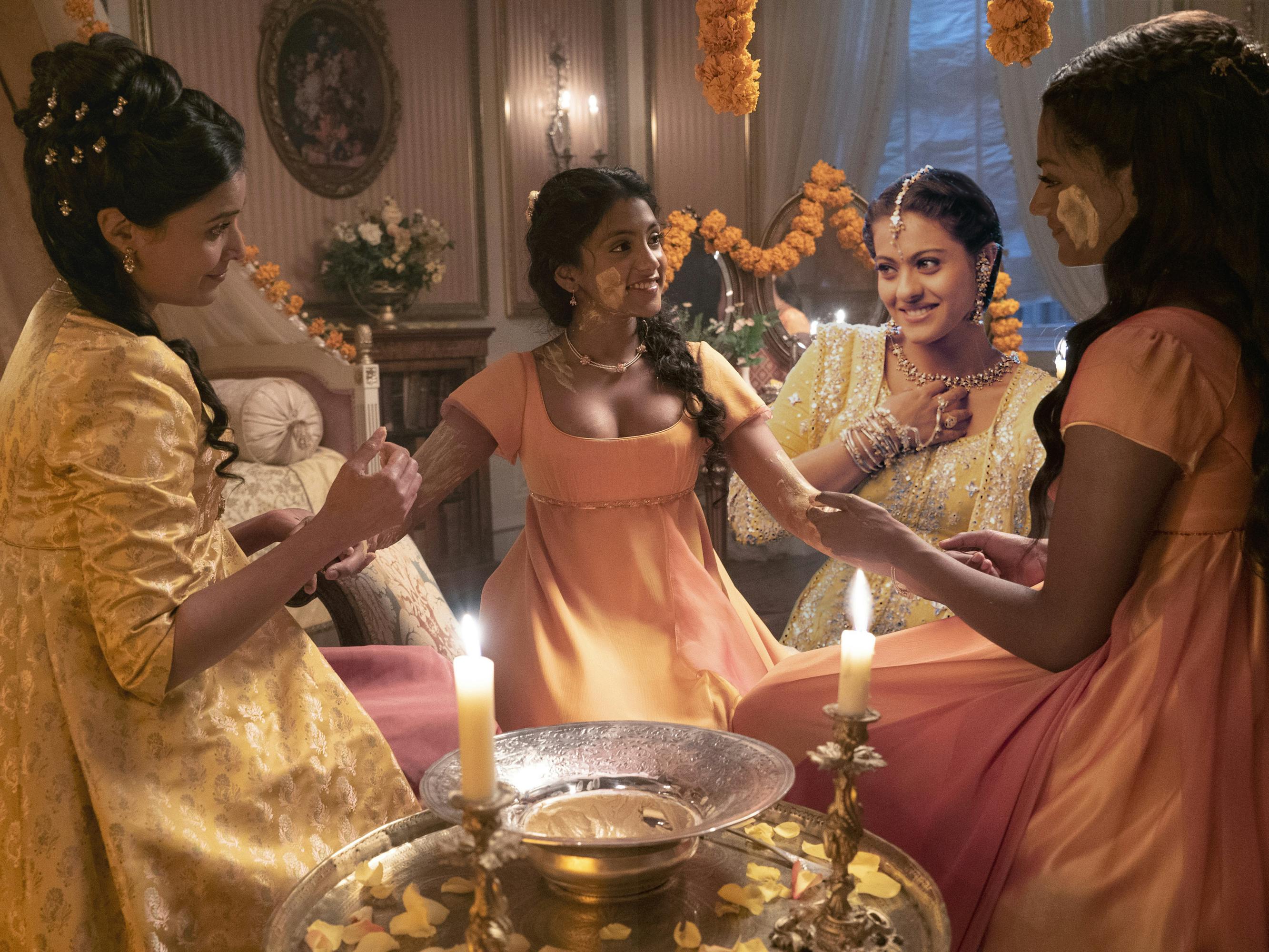 Lady Mary Sharma (Shelley Conn), Edwina Sharma (Charithra Chandran), Kajol (Anjali Sharma) from Kabhi Khushi Kabhie Gham, and Kate Sharma (Simone Ashley) sit around in a haldi ceremony. The Sharma sisters wear orange, and Mary Sharma wears yellow.