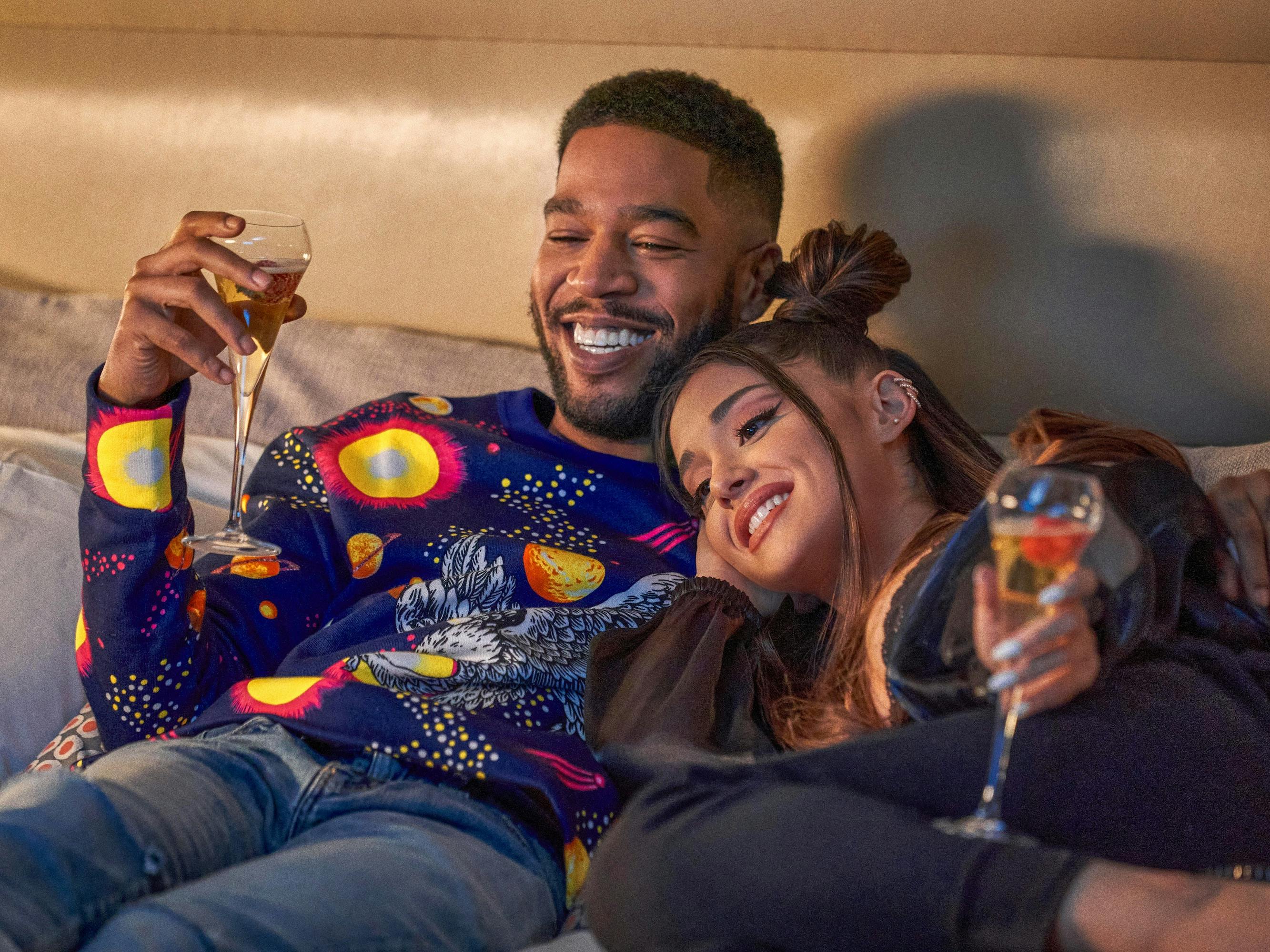 Scott Mescudi and Ariana Grande sit on a couch together drinking fruity champagne. Mescudi wears a patterned sweater and jeans. Grande wears a grey sweatsuit.