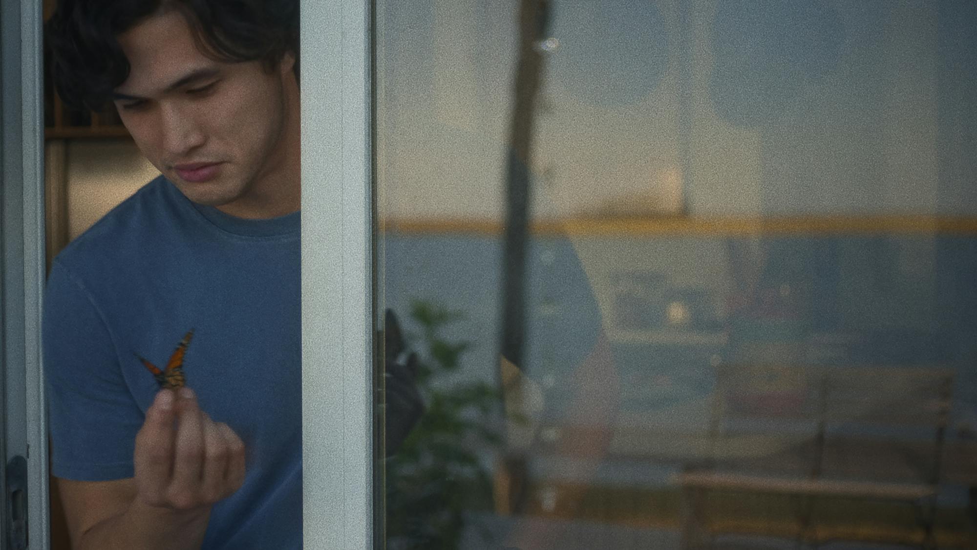 Joe Atherton-Yoo (Charles Melton) wears a blue t-shirt and holds a butterfly out the sliding glass door.