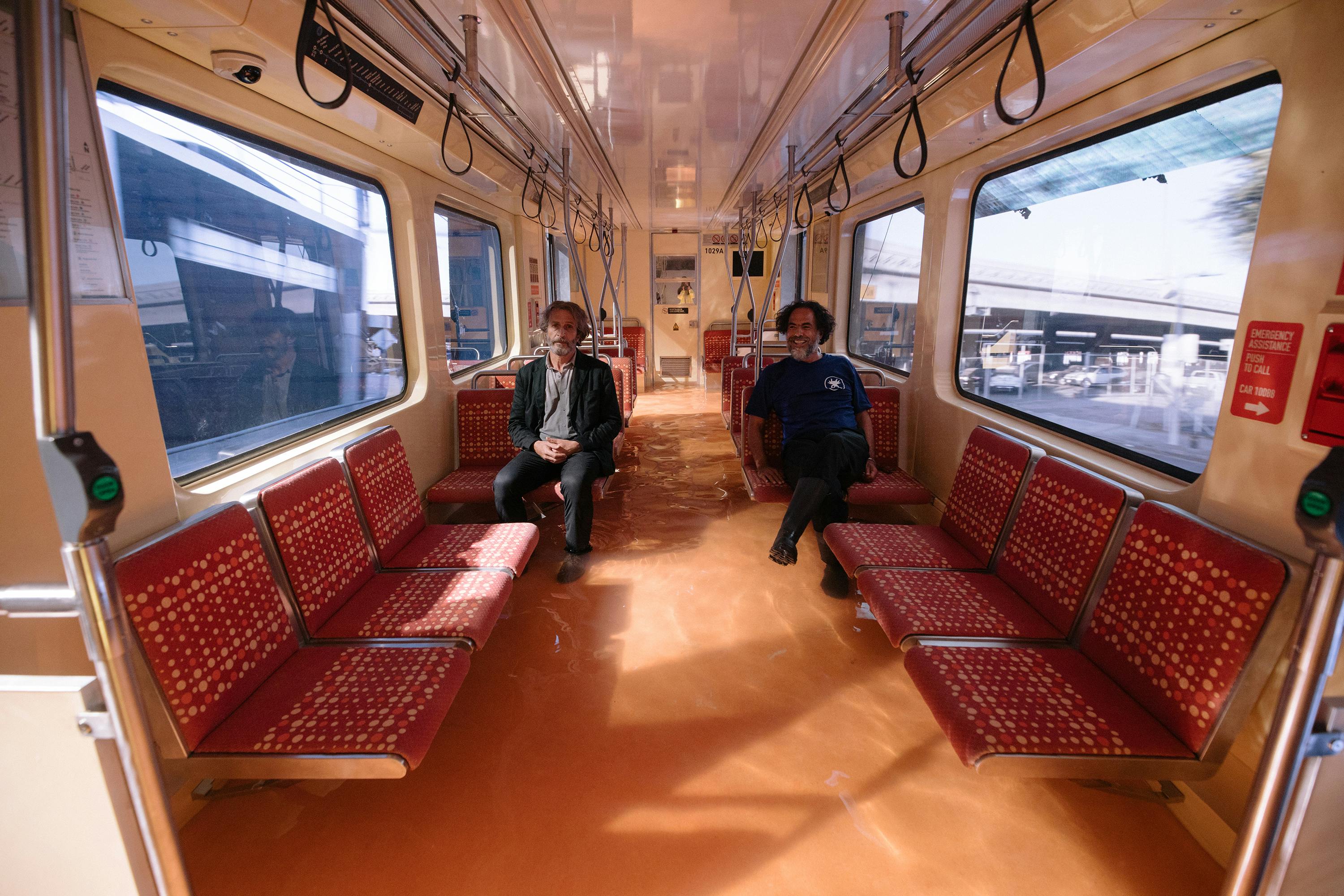 Daniel Giménez Cacho and Alejandro G. Iñárritu sit on a bus together. The bus is lined with red cushioned benches.
