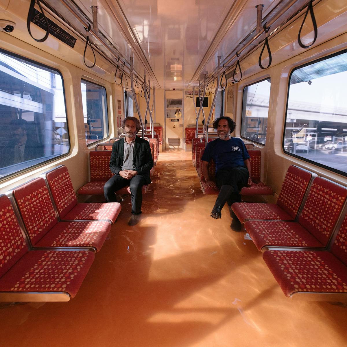 Daniel Giménez Cacho and Alejandro G. Iñárritu sit in a bus lined with red cushioned benches.