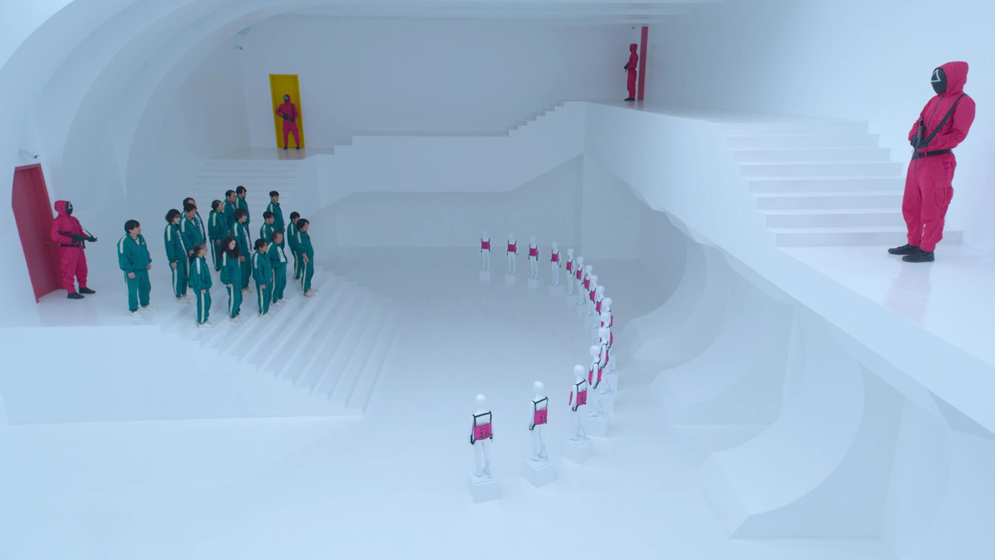 A cluster of green-tracksuit bearing players face off with 16 mannequins wearing pink vests with numbers on the chest. Four pink guards are interspersed throughout the stark white room. 