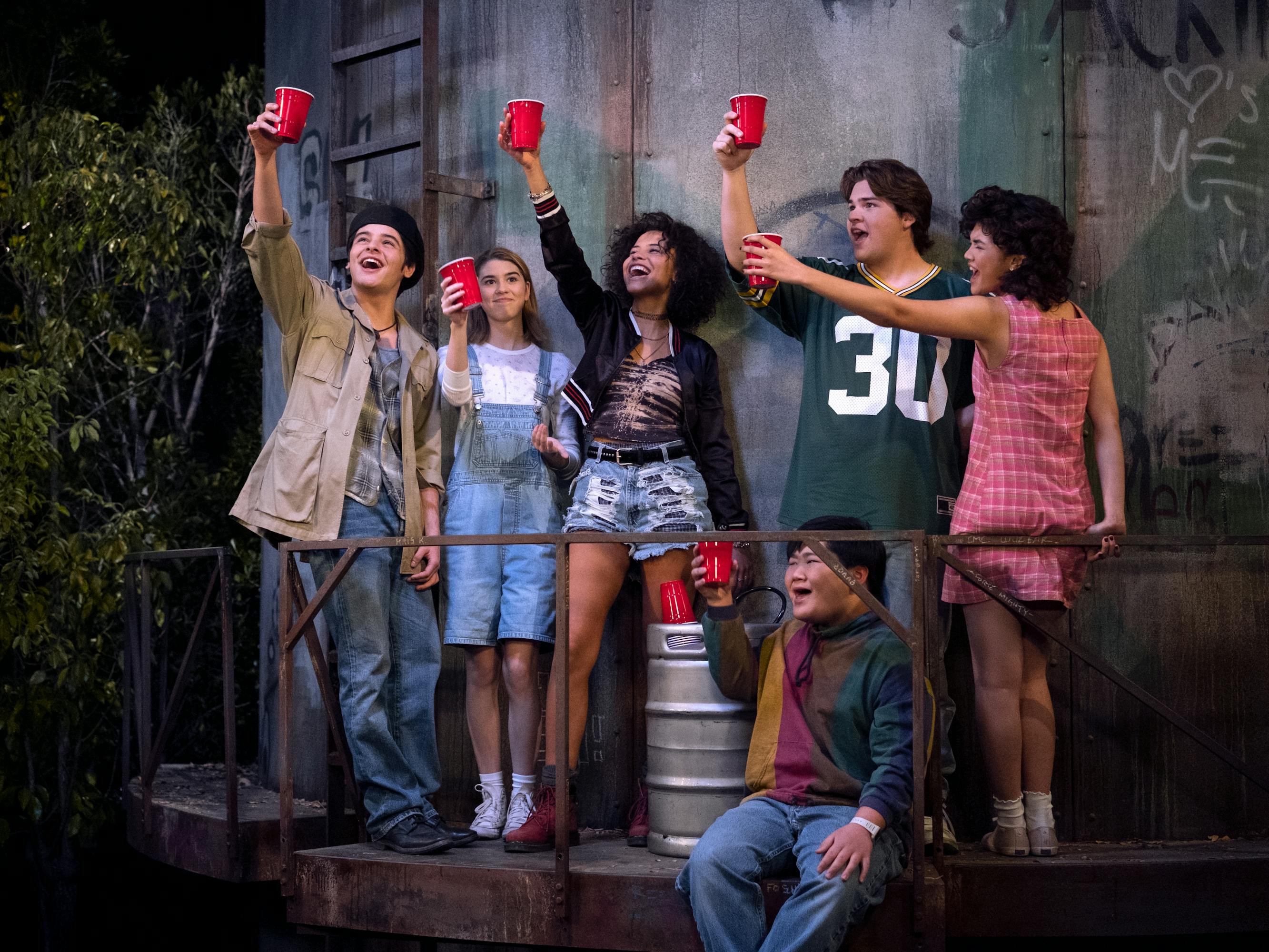 Jay (Mace Coronel), Leia Forman (Callie Haverda), Gwen (Ashley Aufderheide), Ozzie (Reyn Doi), Nate (Maxwell Acee Donovan), and Nikki (Sam Morelos) stand together on a balcony cheersing the sky next to a keg.  