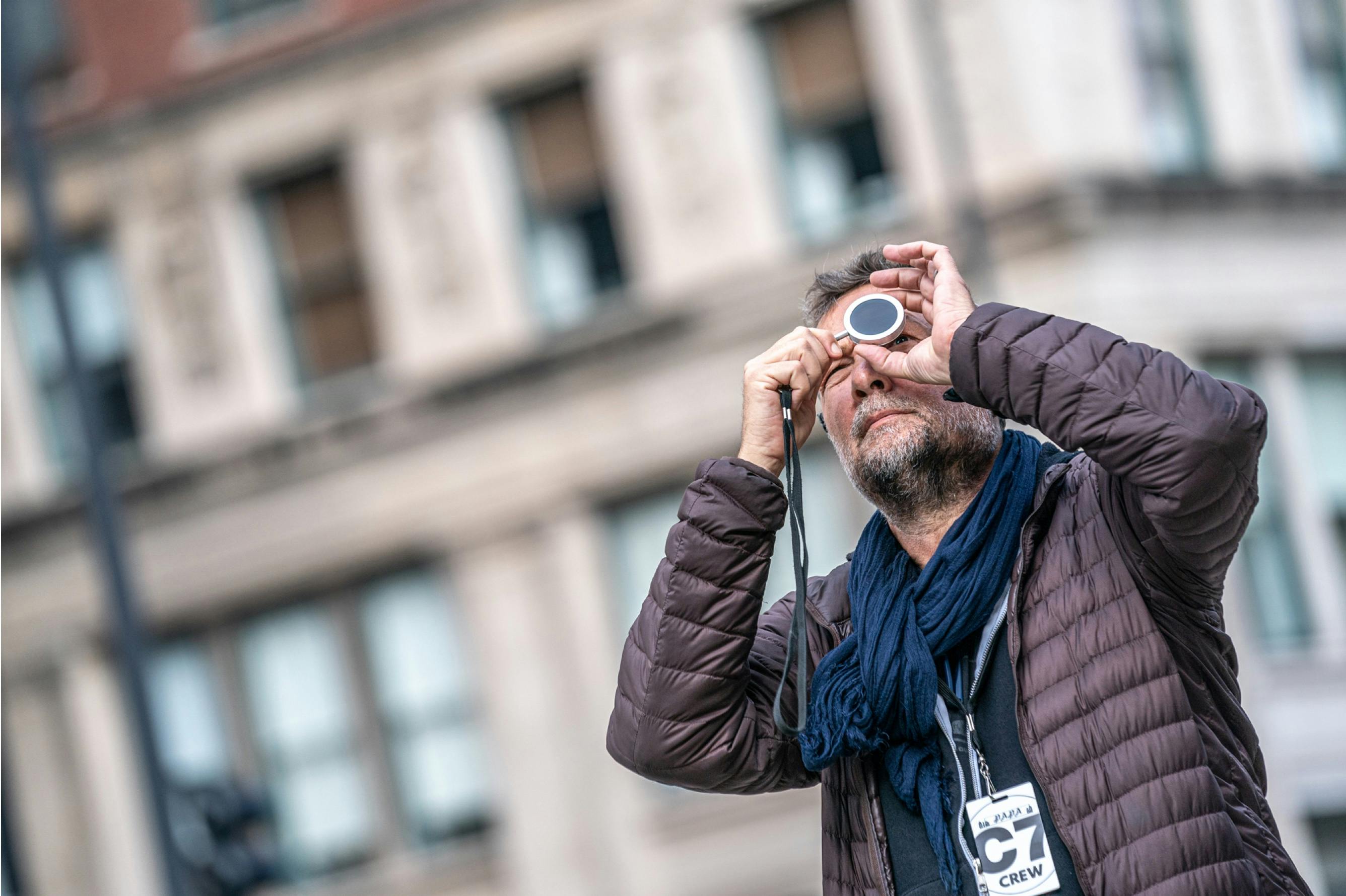 Phedon Papamichael stands on a Chicago street and looks through a lens toward a point above him. There’s a lanyard around his neck with a card that reads, “C7 Crew.” 