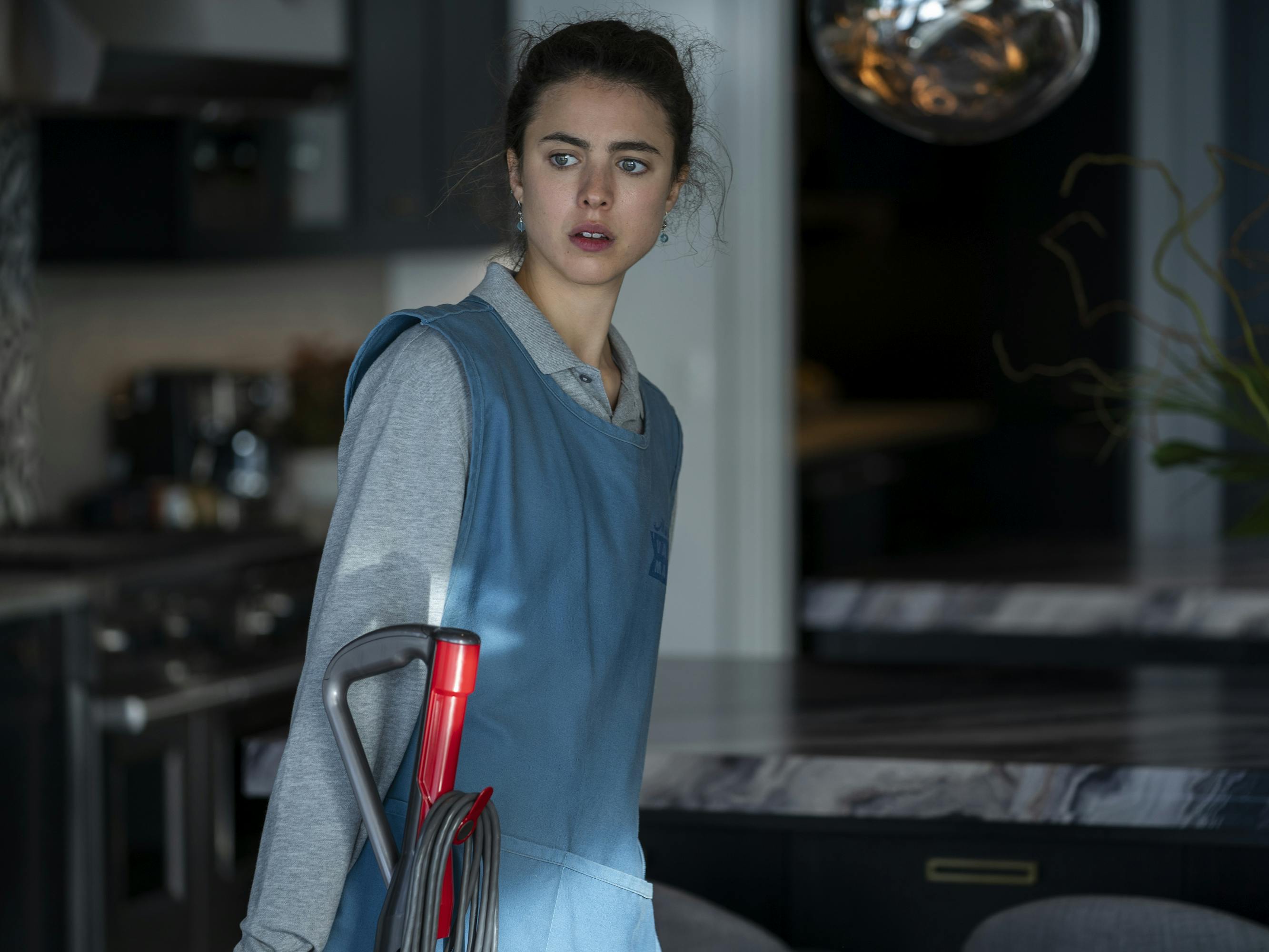 Alex Russell (Margaret Qualley) wears a maid uniform and carries a vacuum. 