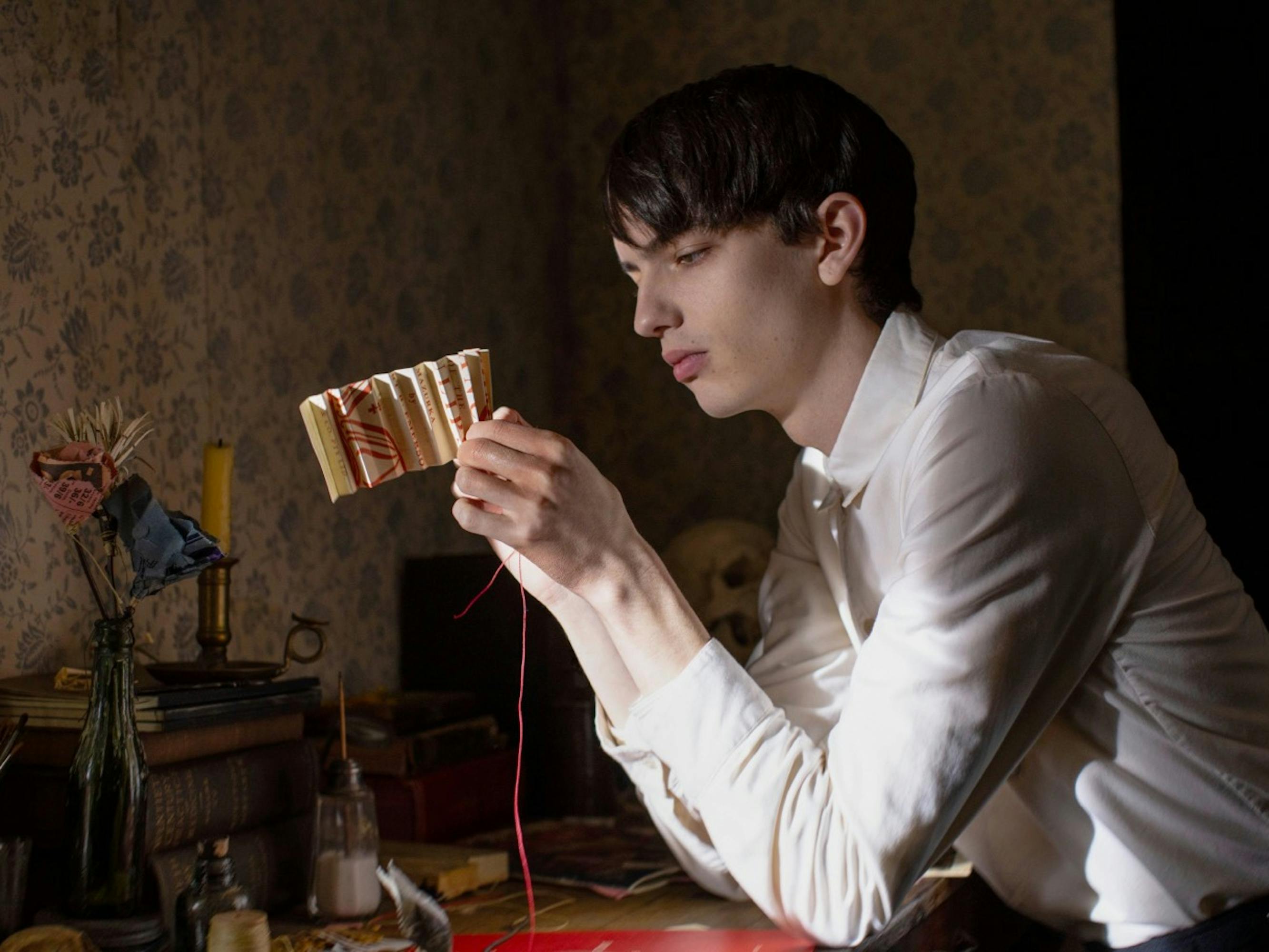 Kodi Smit-McPhee’s character Peter sits alone in a dimly lit room crafting a piece of paper and red string. He wears a white buttoned-down shirt and looks serious.