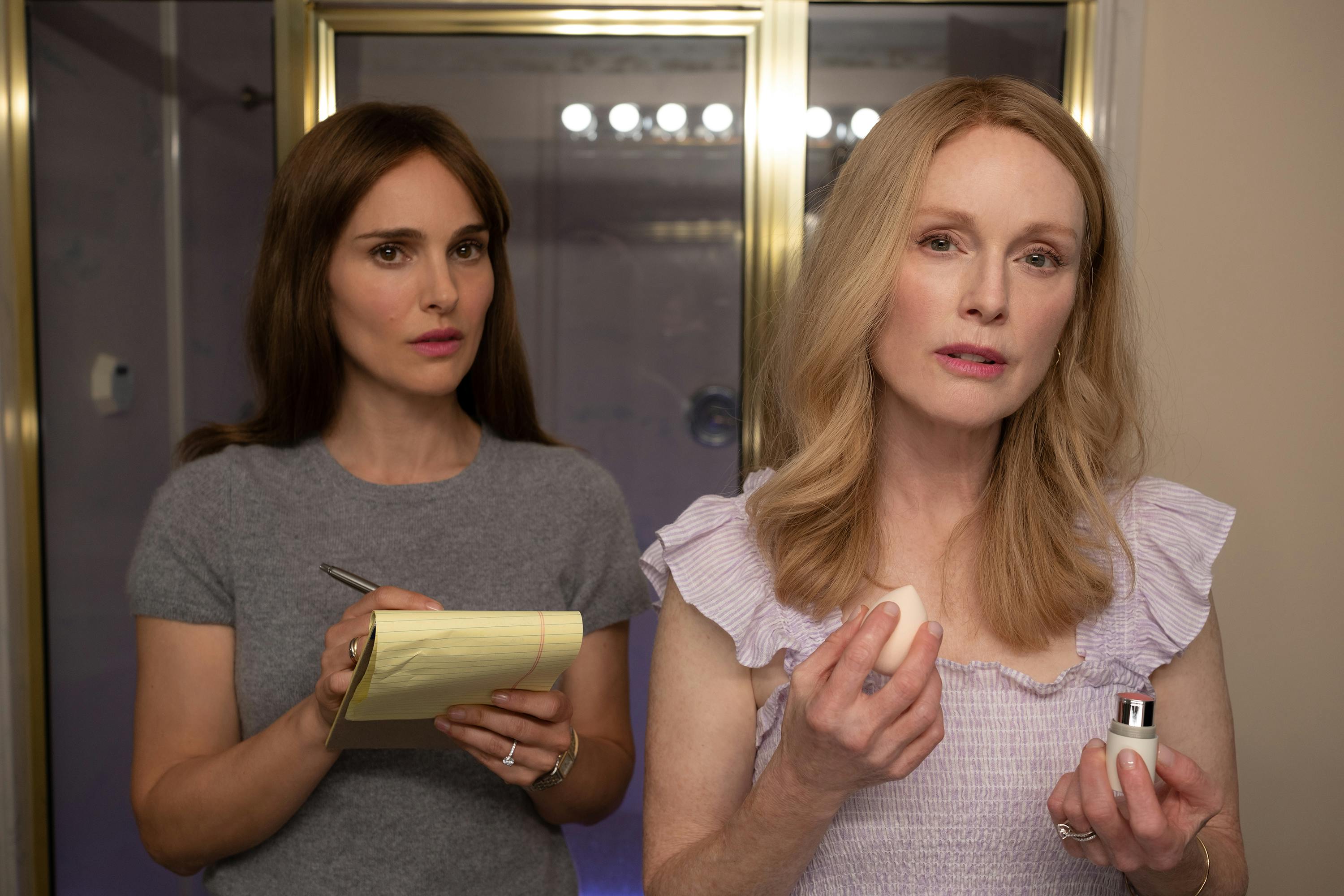 Elizabeth (Natalie Portman) and Gracie (Julianne Moore) stand in front of a lit mirror. Portman wears a grey T-shirt and writes on a small yellow legal pad. Gracie holds a beauty blender and a stick of blush and wears a frilly purple dress.