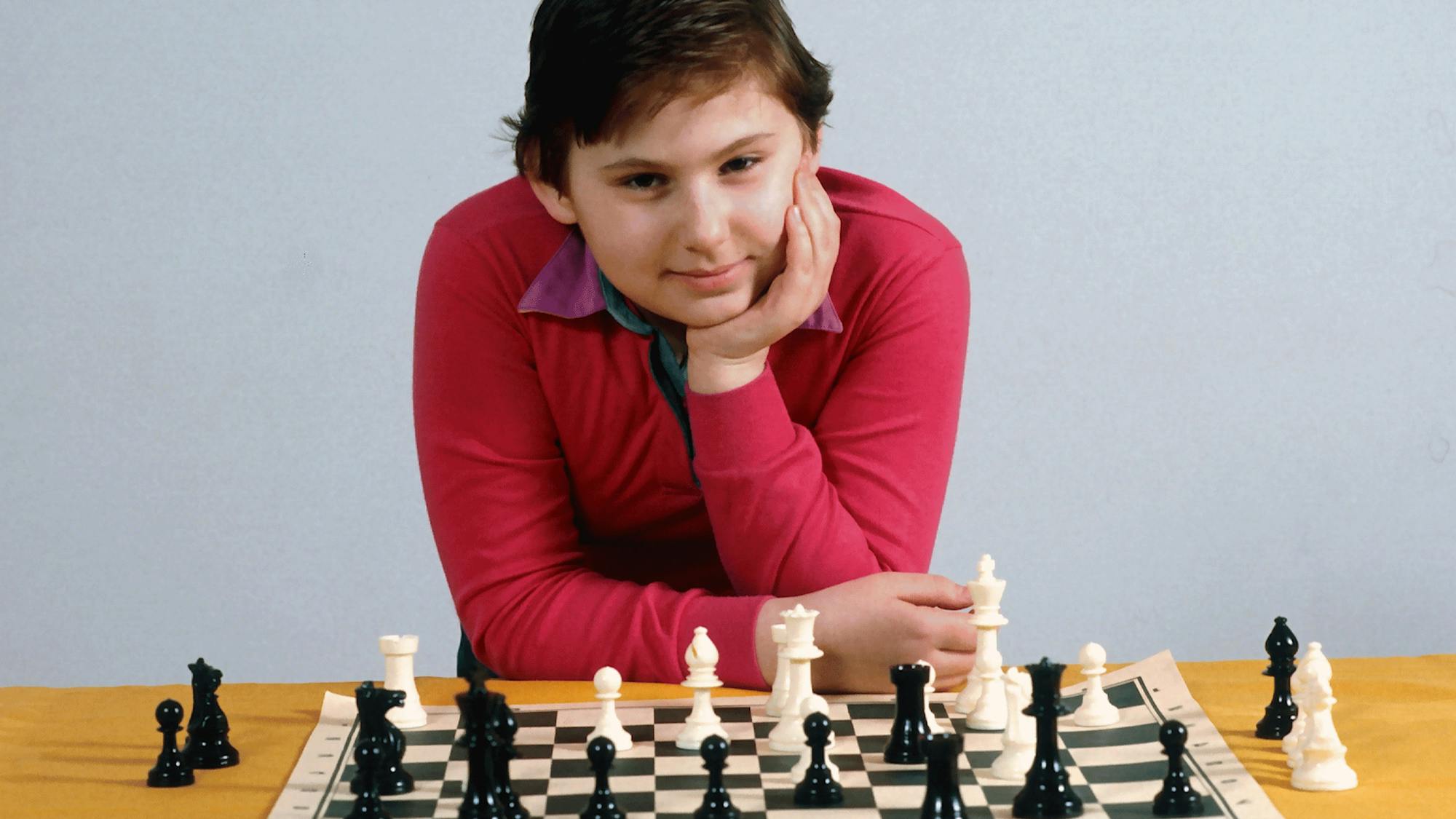 Polgár at age 9. Sporting a bright red shirt and close-cropped hair, she leans over a paper chess board, cupping her face in her hand.