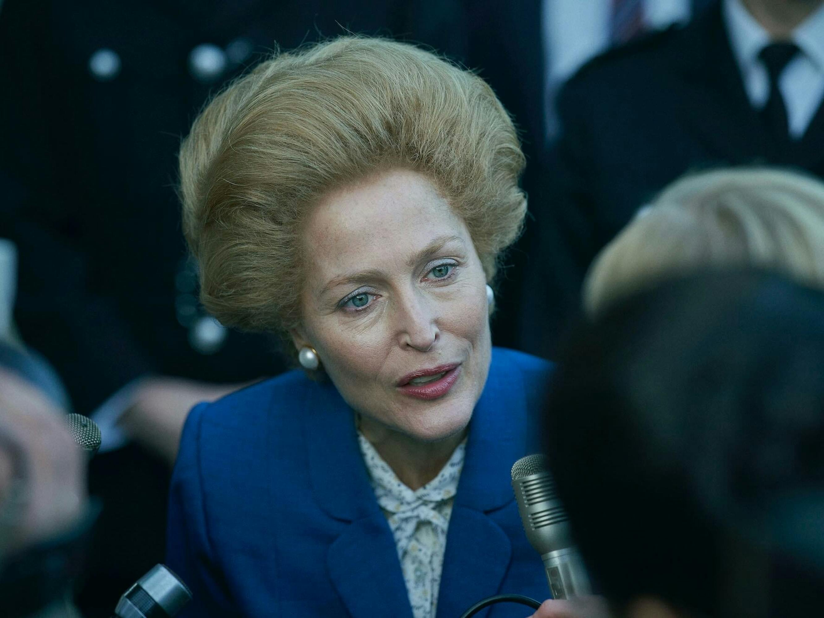 Margaret Thatcher (Gillian Anderson) speaks to reporters. Her blue eyes match her blazer perfectly, and her hair coiffed. Her pearl earrings swallow her ear lobes.