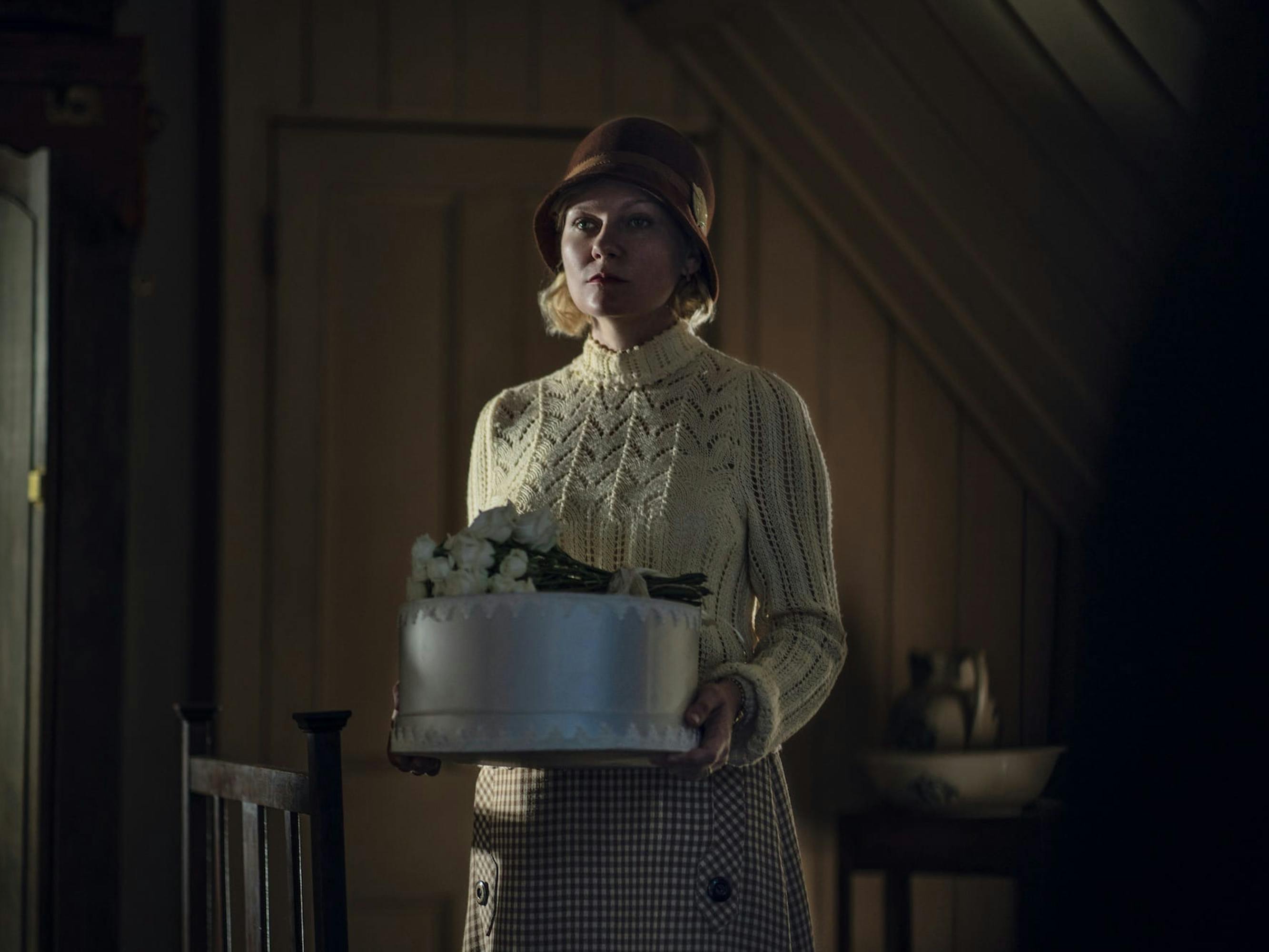 Rose (Kirsten Dunst) carries a white cake, with flowers on top. She wears a white sweater, brown hat, and plaid skirt.