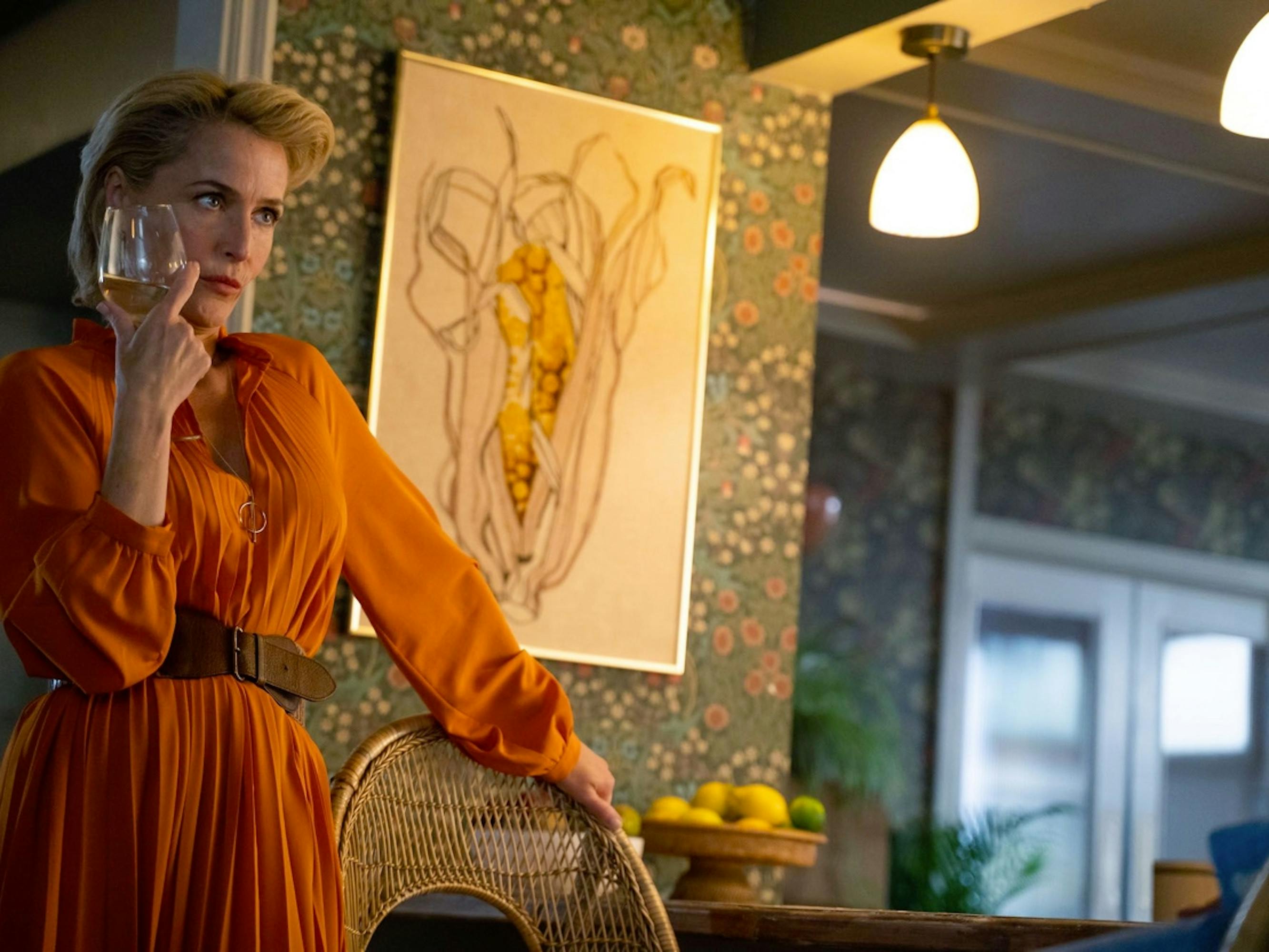 Jean Milburn (Gillian Anderson) presses a glass of chardonnay to her face as she stands in her kitchen. The walls featured a flowery wallpaper, and there is a hanging painting that closely resembles a vagina.