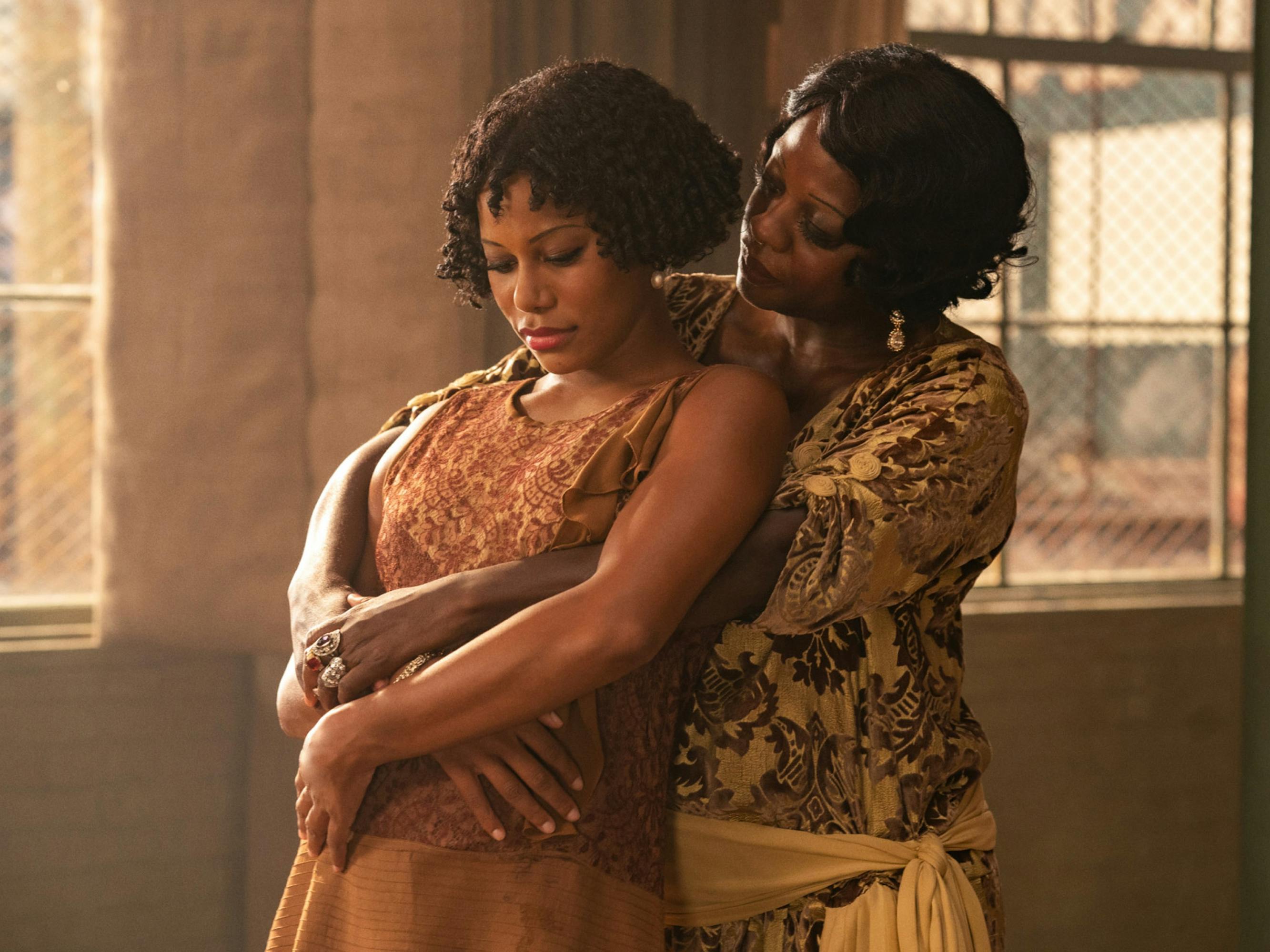Ma Rainey holds her lover Dussie Mae in her arms. She looks attentively at her while Dussie Mae looks down coyly. Warm light pours in from two windows behind them. Dussie Mae wears a chestnut brown dress while Rainey is in golden.