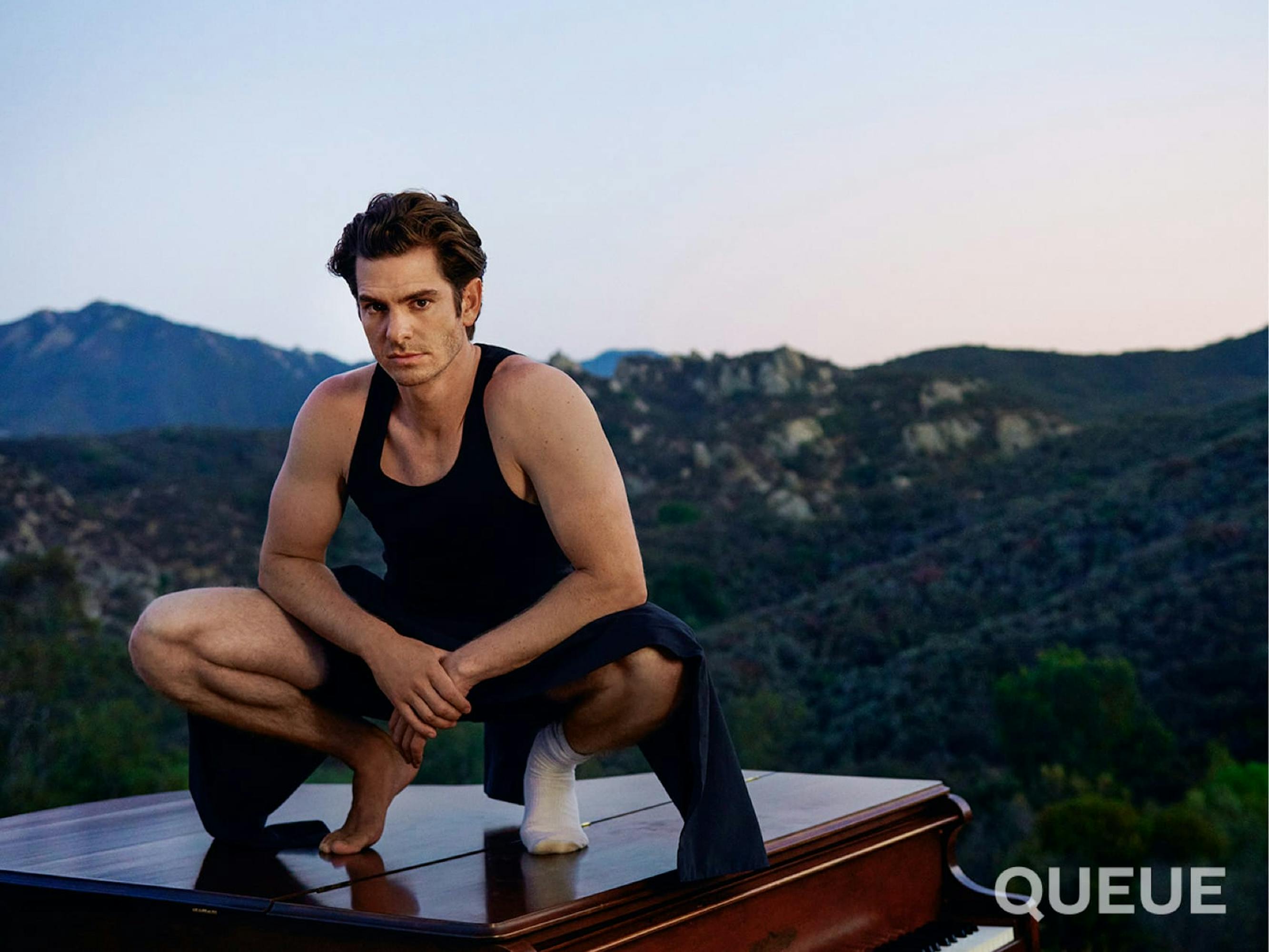 Andrew Garfield wears a dark tank top, dark shorts, and a dark sweater pulled around his waist. On one foot he wears a white sock, and the other is bare. He crouches on a piano, with a magnificent blue-green mountain range in the background, and a pink and blue evening sky.