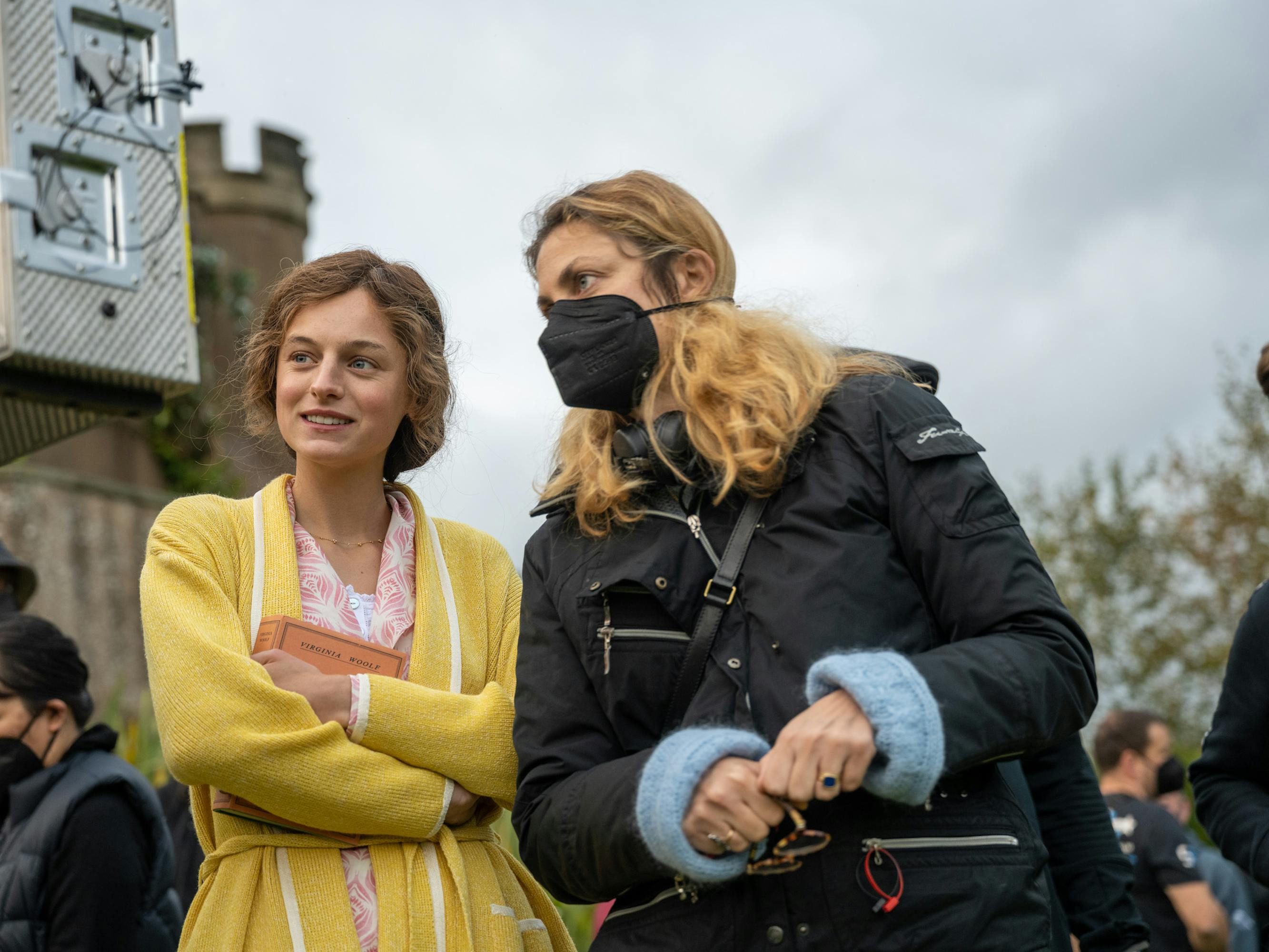 Connie (Emma Corrin) and Laure de Clermont-Tonnerre stand together on set. Corrin wears a yellow robe and Clermont-Tonnerre wears a black puffer.