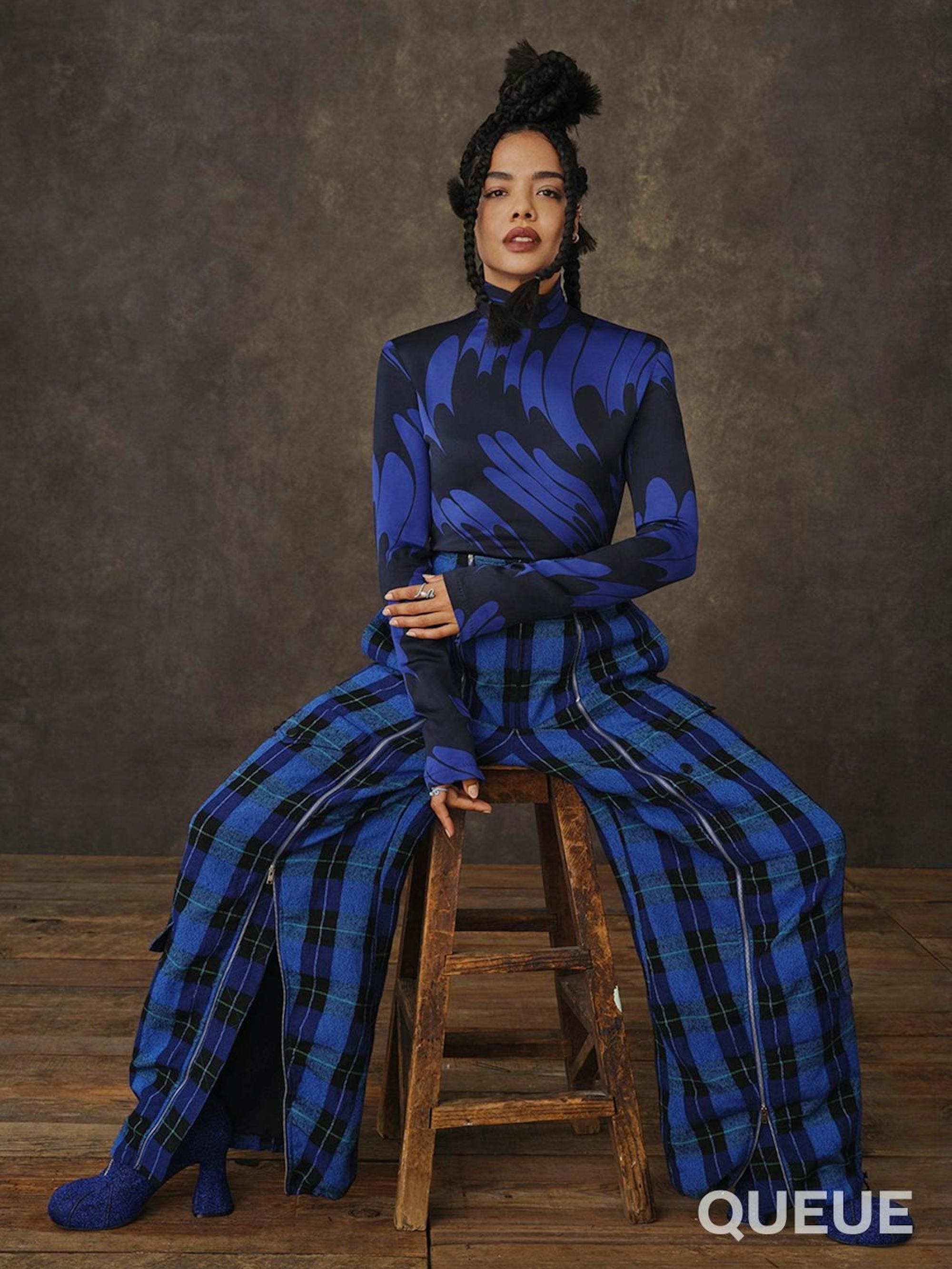 Tessa Thompson wears a blue and black patterned long sleeved turtle neck, and blue-green-black plaid pants. Blue heeled boots peep out from beneath her pants.