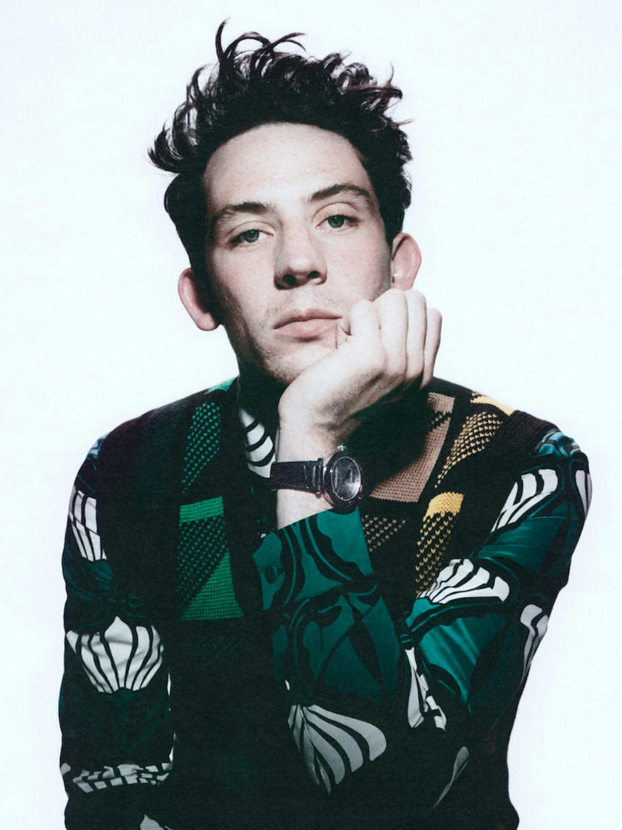 Josh O'Connor sits with his chin resting on the palm of his clenched hand. He wears a white and green floral patterned shirt with a green and yellow wool vest