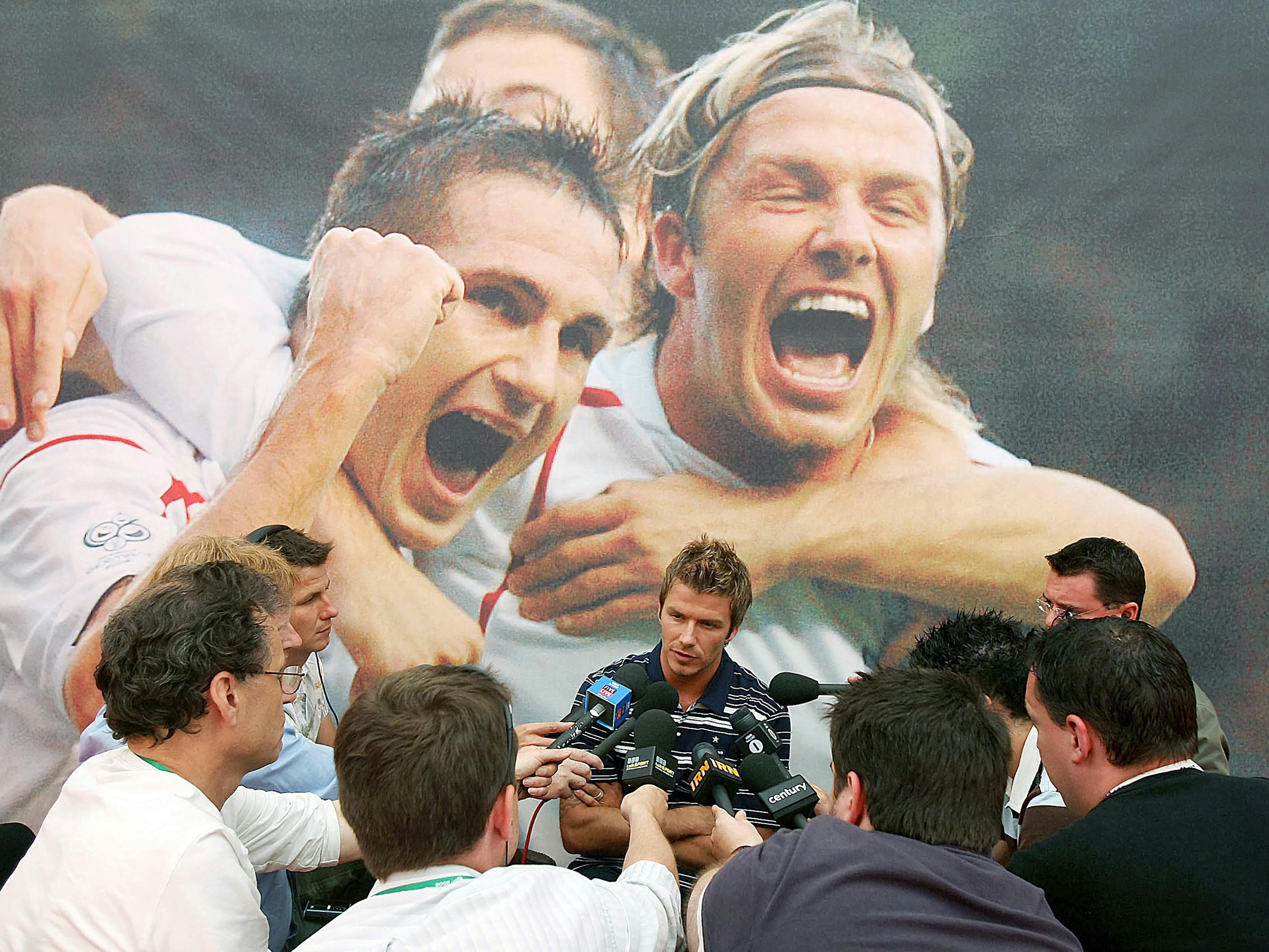 David Beckham gives a press conference to a swarm of reporters, against a backdrop with his and his teammates’ excited faces.