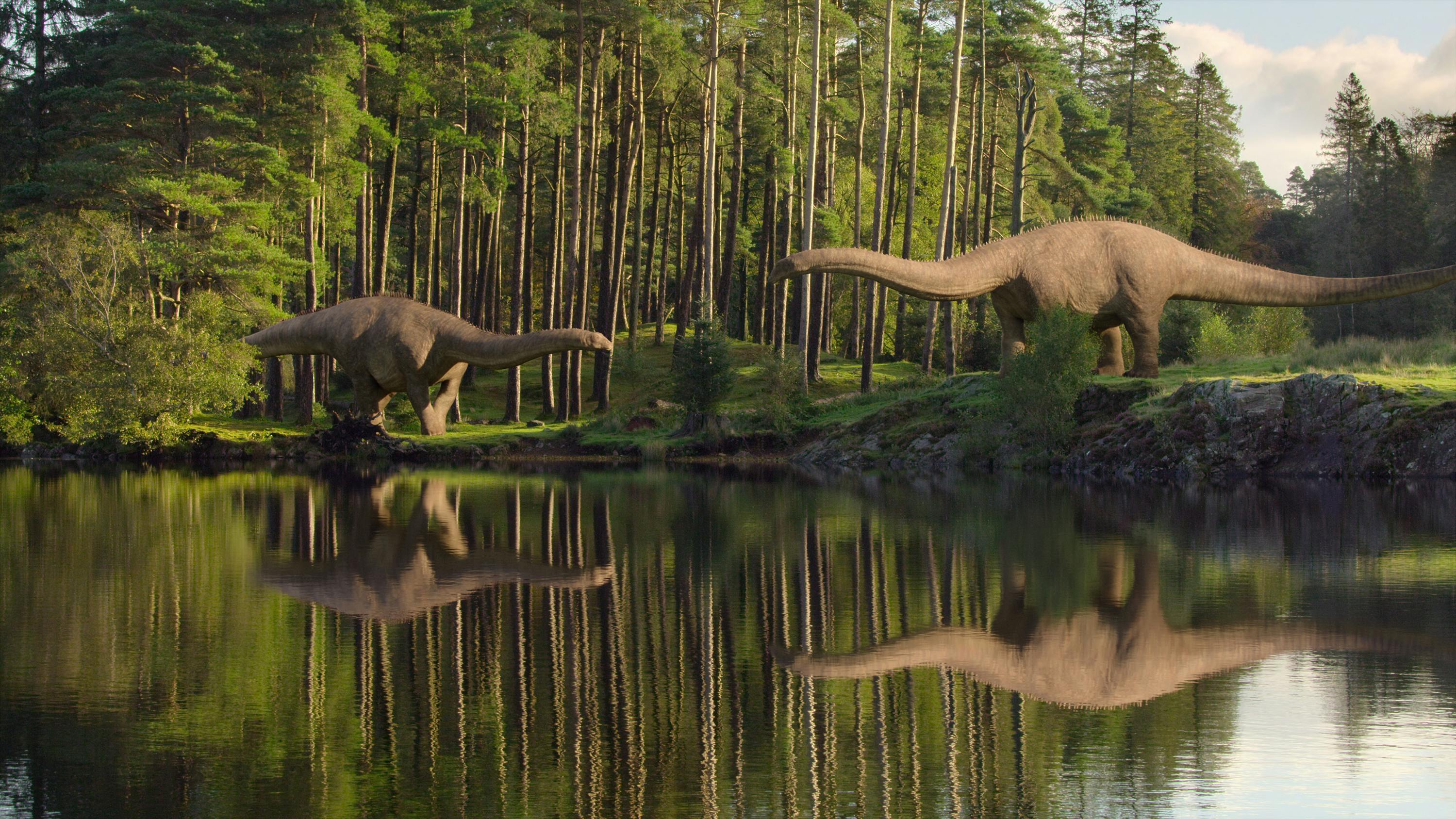 Two dinosaurs stand by a still body of water in a verdant green clearing.