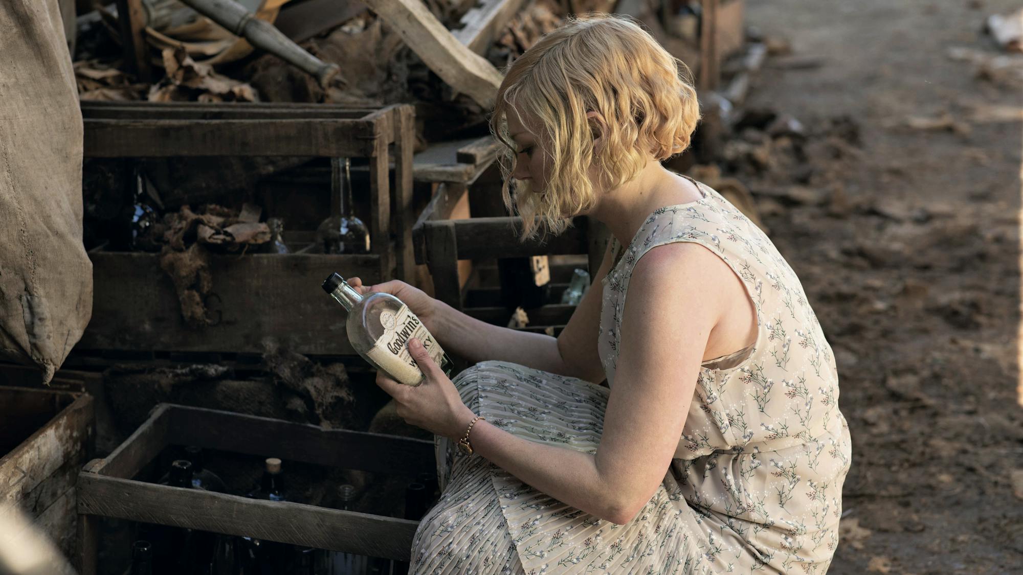 Kirsten Dunst wears a white sleeveless dress and examines a bottle of whiskey from a crate. The ground is muddy.