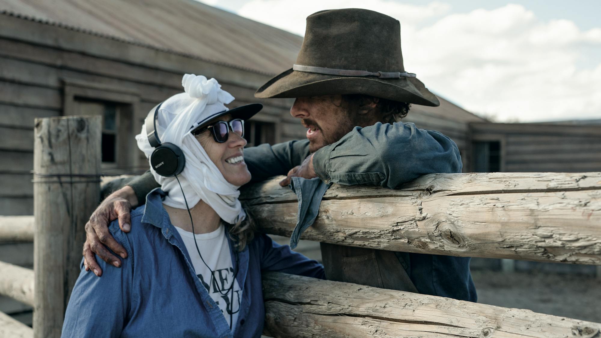 Jane Campion and Benedict Cumberbatch embrace on set on opposite sides of a wooden fence. 