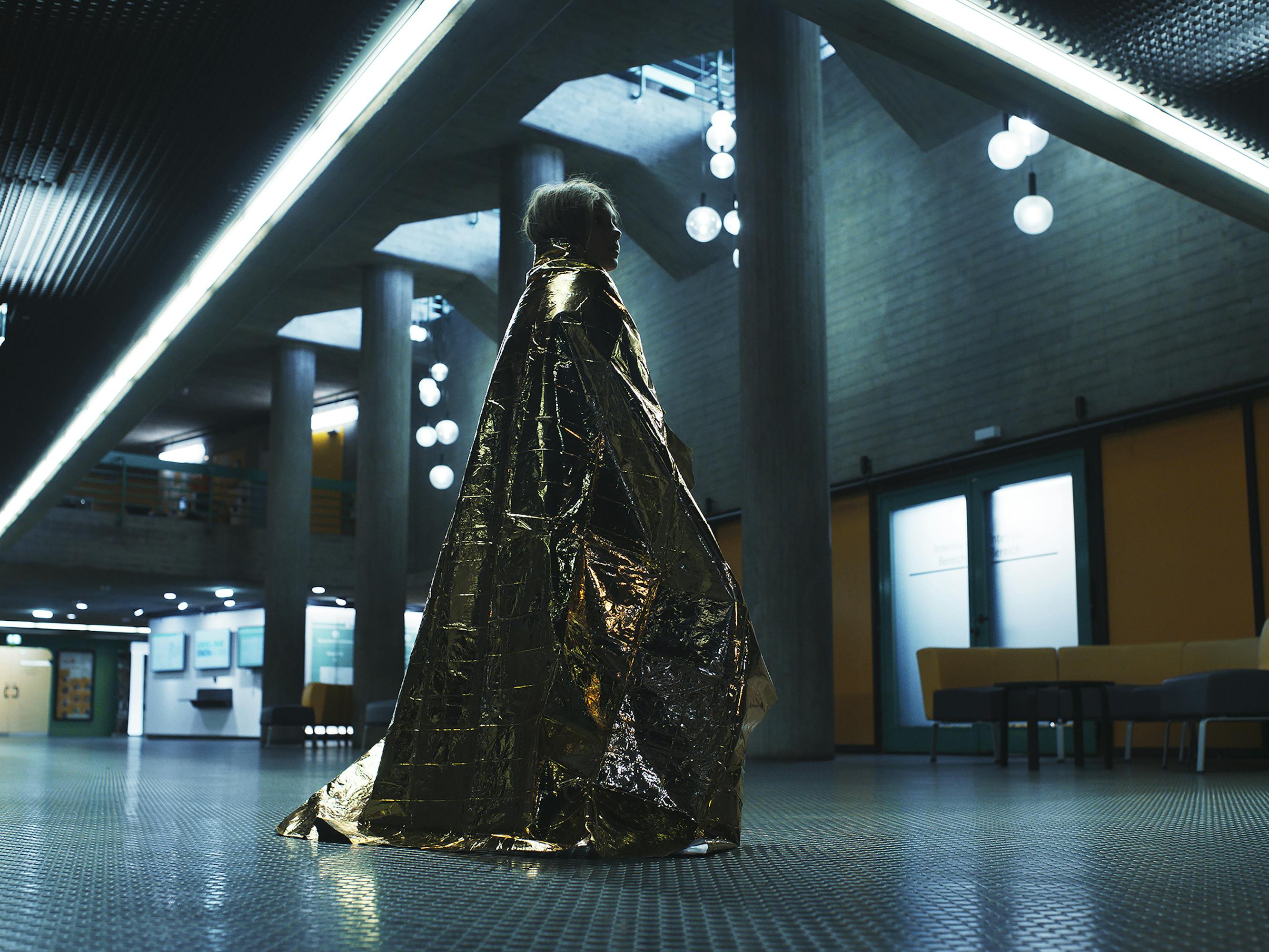 Hannah (Naila Schuberth) wears a gold cloak and walks through a dark hospital. 