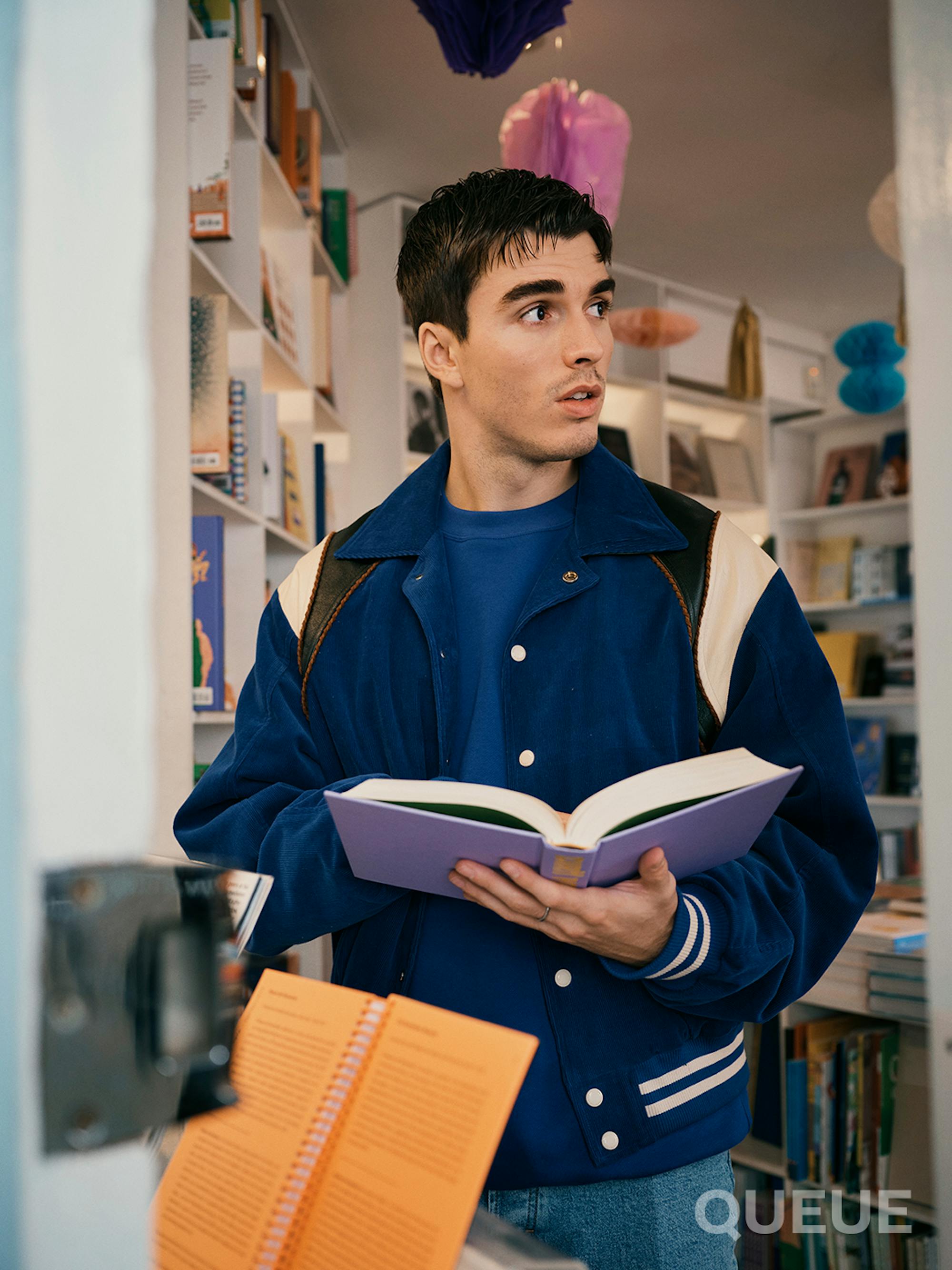 Corey Mylchreest wears jeans, a blue T-shirt, and a blue sports jacket. He holds a book open as he leaves a bookstore.